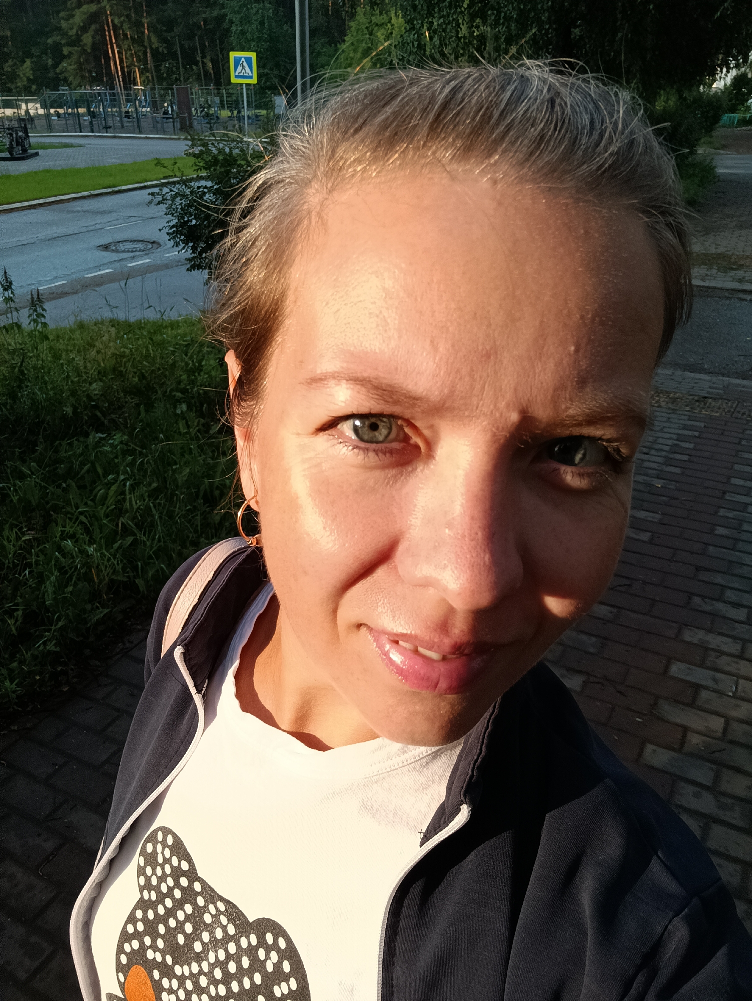 A woman is taking a selfie outdoors, her face close to the camera. She has fair skin, light blue eyes, and her hair is pulled back with some strands falling forward. She is wearing a white t-shirt with a black and white spotted bear design and a dark blue hooded jacket. Her left ear is visible with a gold hoop earring. The background shows a paved walkway, green bushes, a road, and a street sign with a pedestrian crossing symbol. A sports complex with metal structures and trees are visible in the distance. The image appears to be taken during daylight, with sunlight illuminating the woman's face. The overall impression is a candid, outdoor portrait.