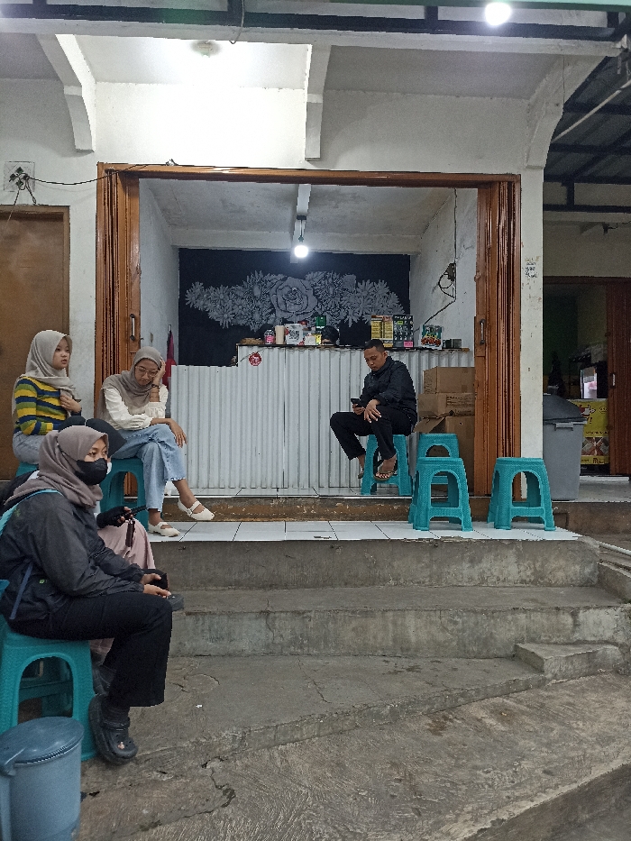 The scene is a small storefront with a concrete patio in front of it. There are four people, three women and one man. Two of the women are wearing headscarves. The man is wearing a black shirt and pants and is sitting on a turquoise plastic chair. The women are sitting on chairs. The man is looking at his phone. The women are looking at the man. The storefront has a large black and white mural of flowers and a sign that says "TOKO". There are also several boxes stacked on the floor. The scene is located in Jatinangor, Indonesia at coordinates -6.935, 107.773. There is a concrete ramp leading up to the storefront.