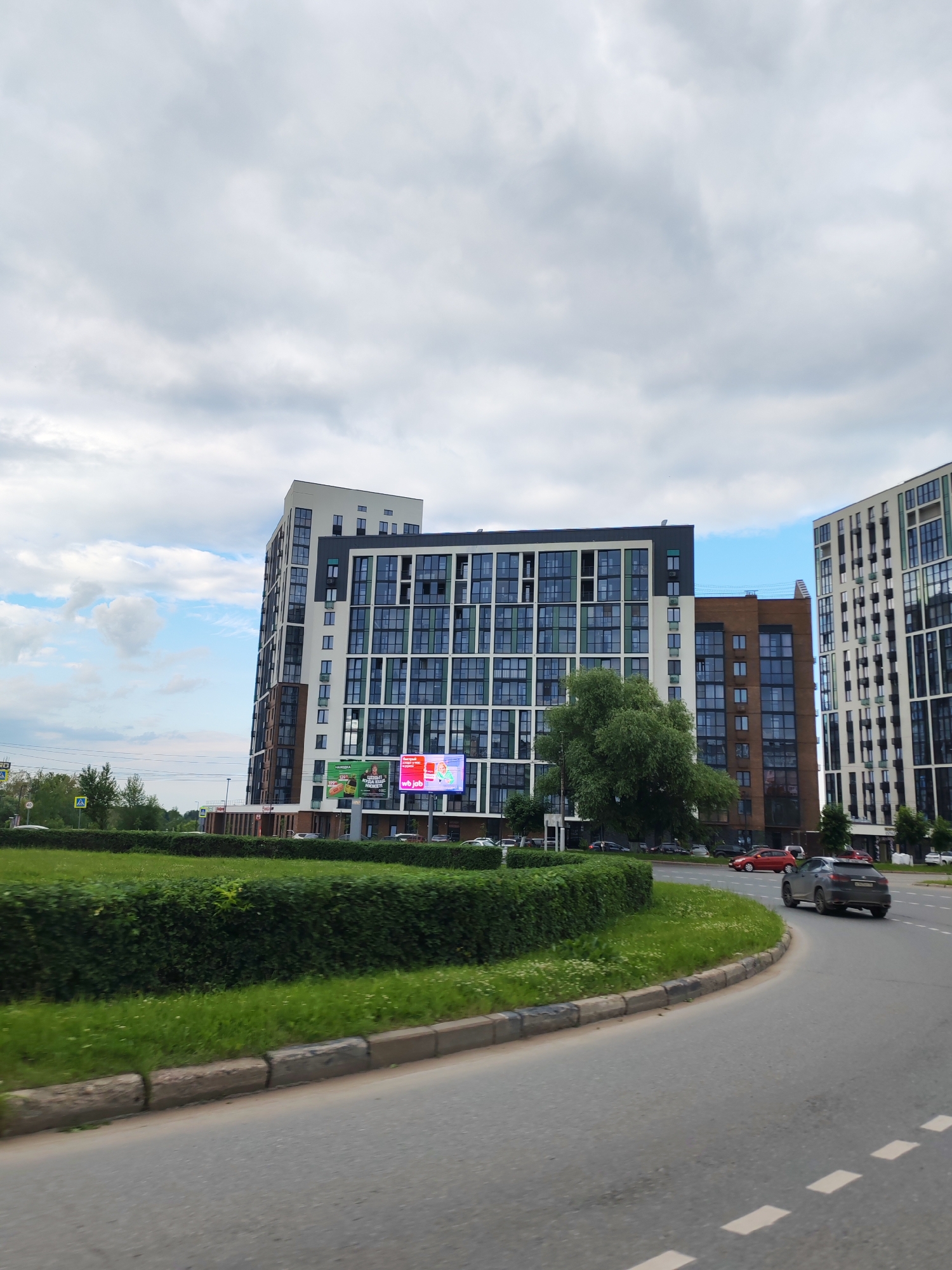 The image shows a view of a modern residential complex in Izhevsk, Russia. The complex features tall buildings with a white and green facade, as well as brown brick sections. The buildings have many windows, giving them a contemporary look. In front of the buildings, there is a neatly trimmed green hedge and a grassy area. A curved road is visible in the foreground, with a dark grey SUV driving on it. The sky is cloudy with some patches of blue. In the background, more buildings and trees can be seen. There are also several billboards on the side of one of the buildings, displaying advertisements.
