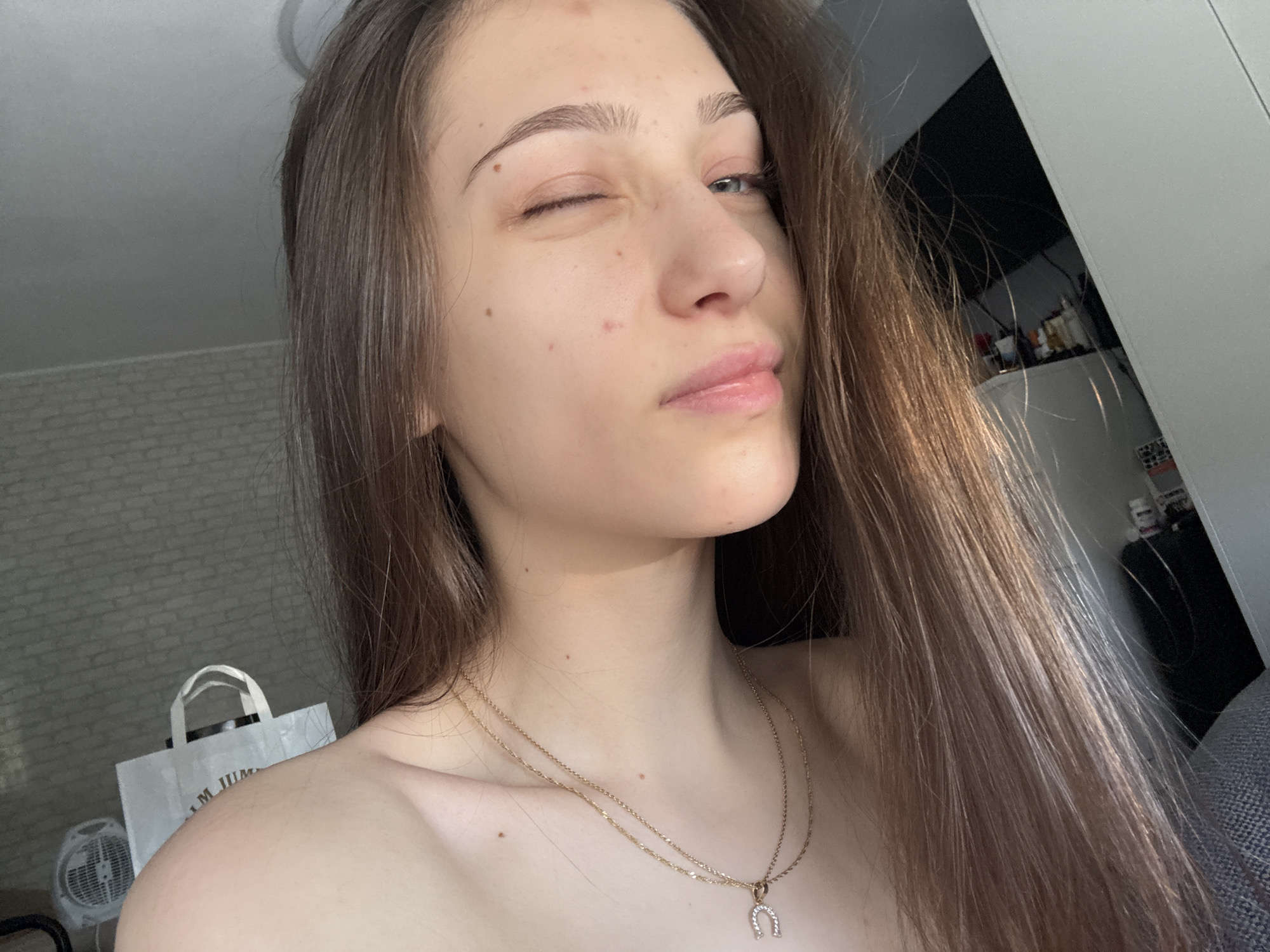 A young woman is taking a selfie indoors. She is winking with her left eye. She has long brown hair, and is wearing a gold necklace with a horseshoe pendant. The background includes a shopping bag, a fan, and a glimpse of a modern interior with white walls and a dark-colored piece of furniture. There is not enough information to know that this photo was taken in Sofia, Bulgaria.