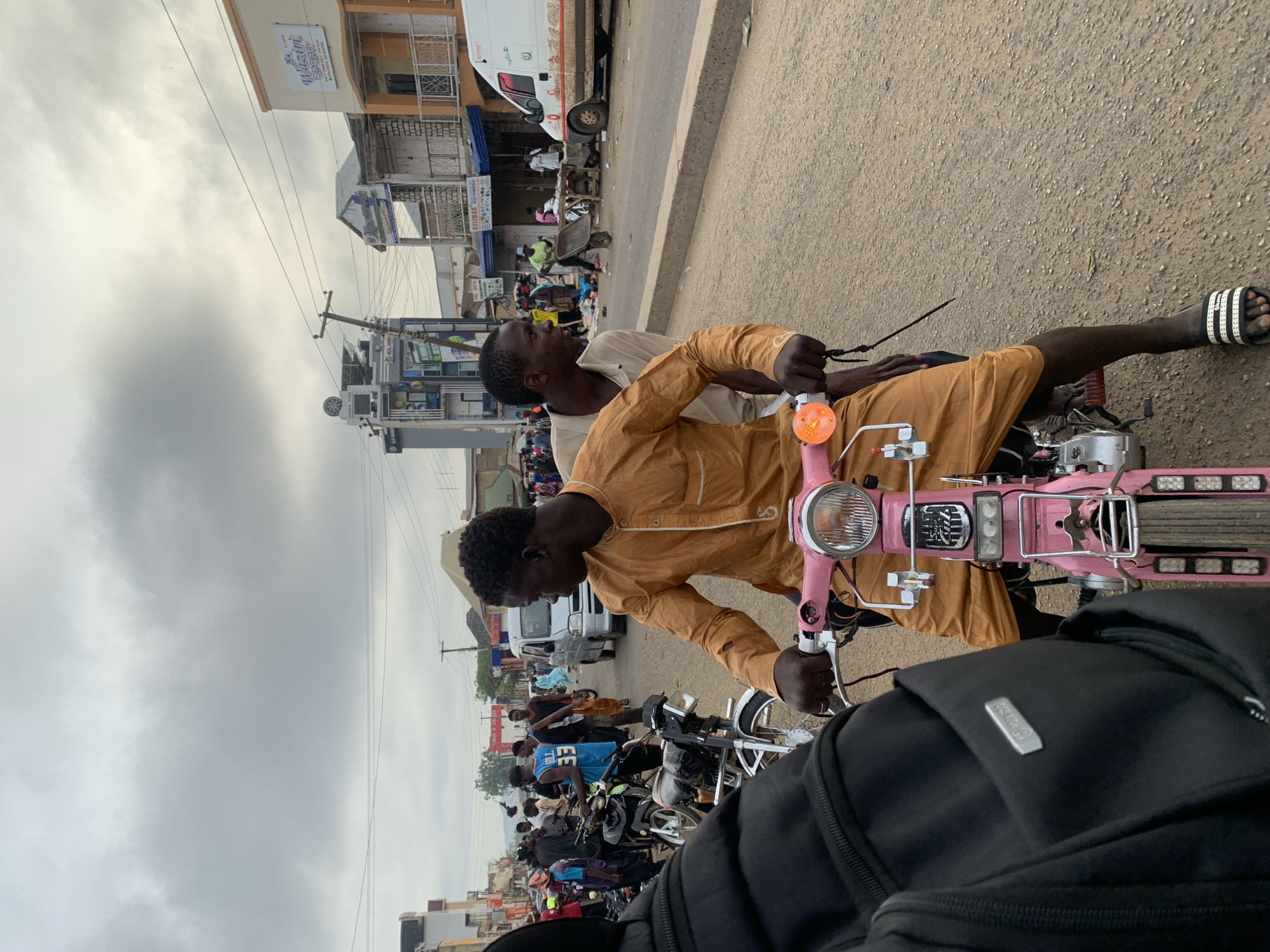 Two men are riding a pink motorcycle on a street in Jos, Nigeria. The driver is wearing a mustard-colored robe and Adidas sandals. The passenger is wearing a light-colored shirt. There are buildings and other vehicles visible in the background. Many people and motorbikes are present, suggesting a busy street scene. Overhead, the sky is overcast. The road is paved.