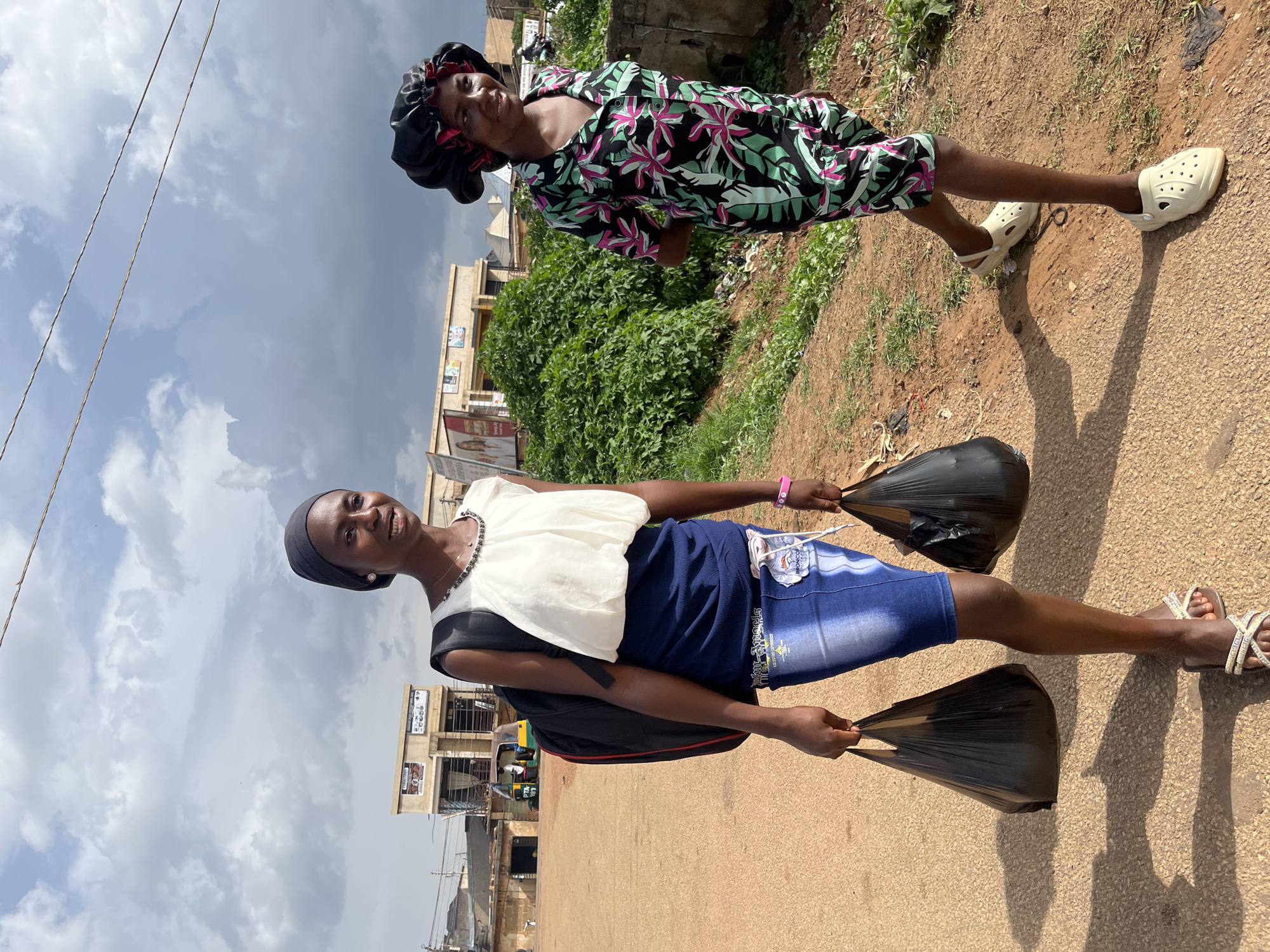 Two young women are walking on a dusty road in Jos, Nigeria. The woman on the left wears a dark headscarf, a white blouse, and a blue skirt. She carries a backpack and two black plastic bags. The woman on the right wears a black satin bonnet and a floral print shirt and shorts. She is walking with her hands behind her back. The background shows buildings, vegetation, and a cloudy sky. A tuk-tuk is visible in the distance.
