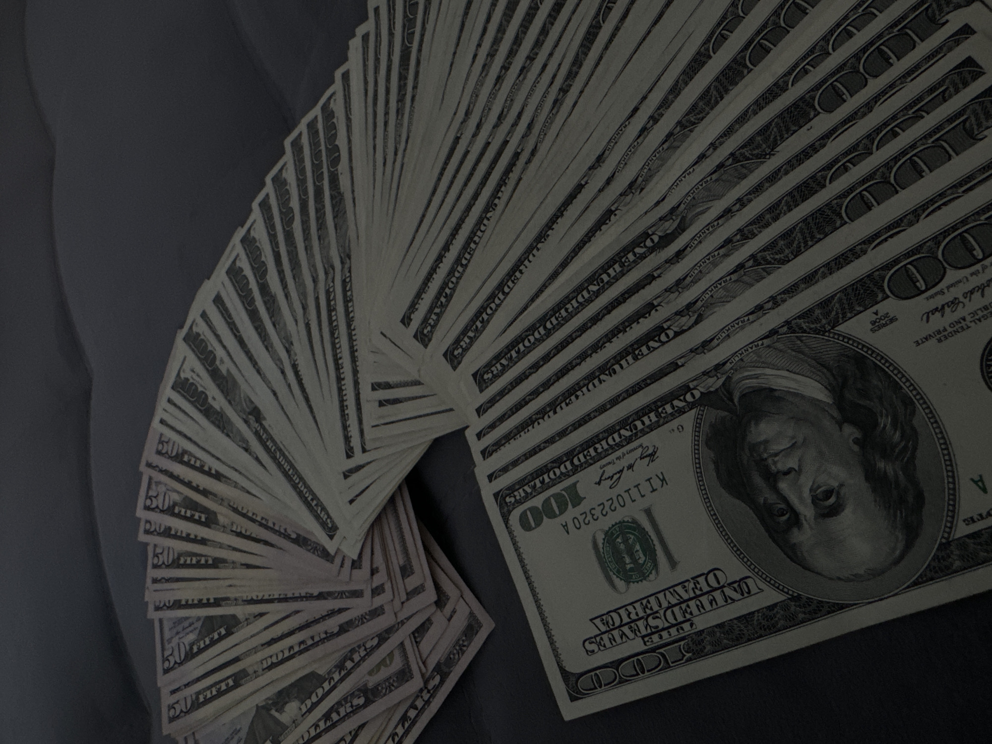 The image shows a fan of US dollar bills, primarily one hundred dollar bills and some fifty dollar bills, fanned out on a dark surface. The bills are spread in a semi-circle, with the one hundred dollar bills fanned out on the right and the fifty dollar bills fanned out on the left, creating a layered effect. The bills are the main subject, and there are no people or discernible scene beyond the bills and the dark surface they are resting on. The lighting appears to be artificial and subdued, casting shadows that add to the low-key aesthetic. There is no indication of time of day, weather, or specific location cues within the image itself. The only visible text is that printed on the US currency, which includes denominations like "ONE HUNDRED DOLLARS" and "FIFTY DOLLARS," as well as portraits and other security features of the bills. The overall impression is one of wealth or financial display.