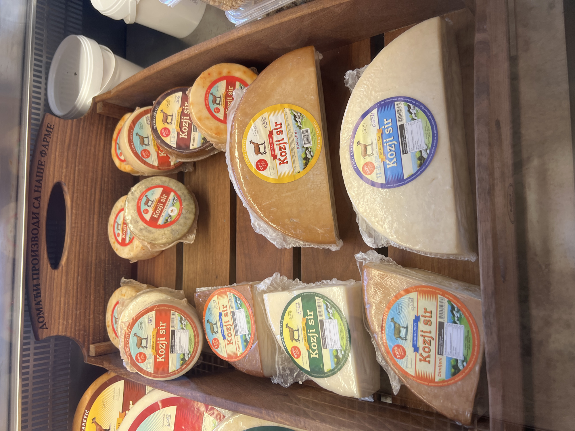 The image shows a close-up view of a display of goat cheese at a farm shop, likely in Kovačko Polje, Montenegro, based on the text. Several wheels and wedges of goat cheese are presented in wooden crates, with clear plastic packaging and labels indicating "Kozji sir" (goat cheese) and "Farma Carevic". The labels also feature graphics of goats and some nutritional information. In the background, white buckets are visible, and a wooden sign with Cyrillic script reading "ДОМАЋИ ПРОИЗВОДИ СА НАШЕ ФАРМЕ" (Homemade products from our farm) is positioned above the cheese display. The lighting suggests an indoor setting, possibly a refrigerated display case.