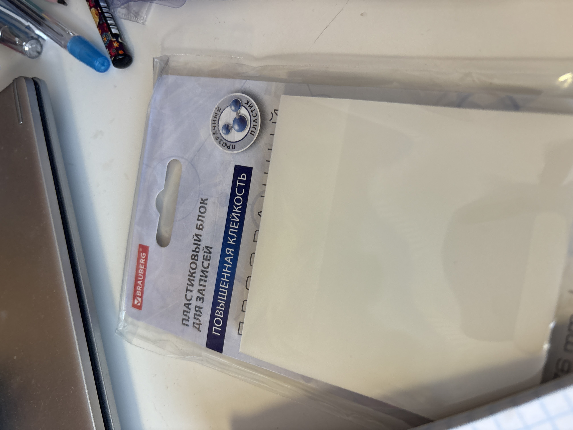 A clear plastic package containing a block of white paper is placed on a light-colored, flat surface. The package has a white and light blue header with Russian text. The text on the header reads "BRAUBERG", "ПЛАСТИКОВЫЙ БЛОК ДЛЯ ЗАПИСЕЙ" (Plastic block for notes), and "ПОВЫШЕННАЯ КЛЕЙКОСТЬ" (Increased adhesiveness). A circular emblem with blue spheres and Russian text "ПРОЗРАЧНЫЙ ПЛАСТИК" (Transparent plastic) is visible on the right side of the header. To the upper right of the package, several pens are visible, including a blue pen and a pen with a colorful, patterned barrel. A portion of a silver laptop is in the upper left corner of the image. The lower left corner shows a grid pattern, likely from a notebook. The overall scene suggests an office or desk setting. The image's context does not provide any information related to the city of Georgiyevsk, Russia.