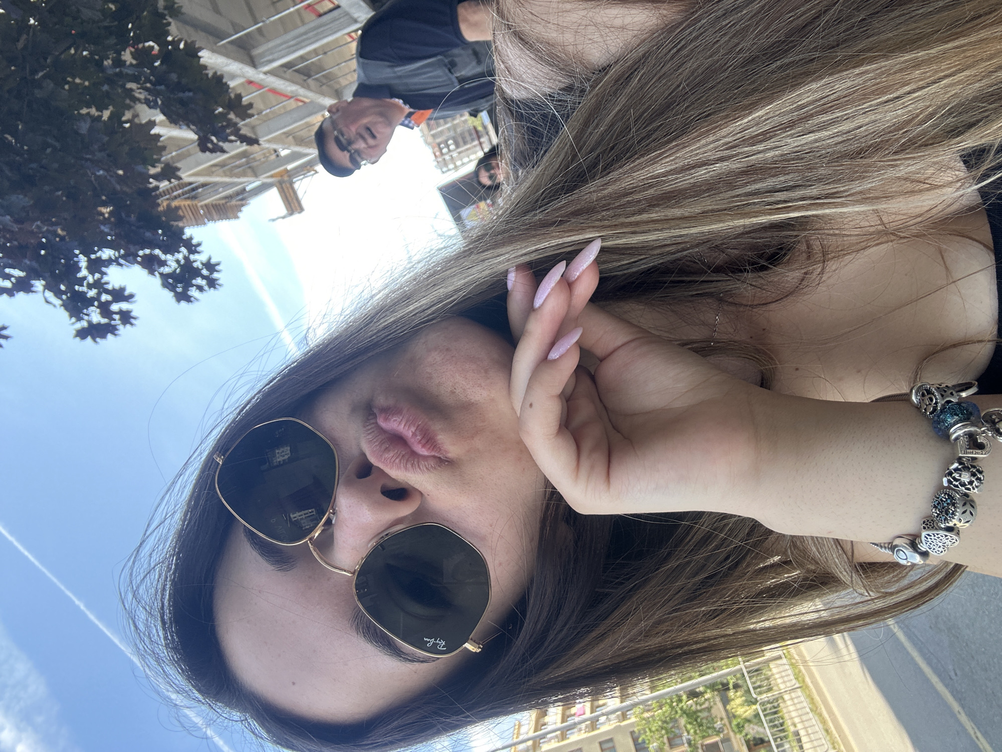 In a selfie taken outdoors, a young woman with long, brown hair and light skin is positioned in the foreground, looking towards the camera with her lips pursed in a kiss pose. She is wearing gold-rimmed sunglasses with dark lenses, and her right hand is resting on her chin, showcasing a bracelet with various charms on her wrist. Her fingernails are painted with a shimmery pink polish.
In the background, an older man with glasses and a dark vest over a light blue shirt is visible, looking towards the right. Behind him, a building under construction with scaffolding is present. The sky is bright blue with a faint white contrail. A tree with dark foliage is partially obscuring the building. The setting appears to be an urban environment, possibly a street, with a white fence and greenery visible on the left side. The overall atmosphere suggests a sunny day in a city. Without further information, it is not possible to definitively identify the specific location in Sofia, Bulgaria.