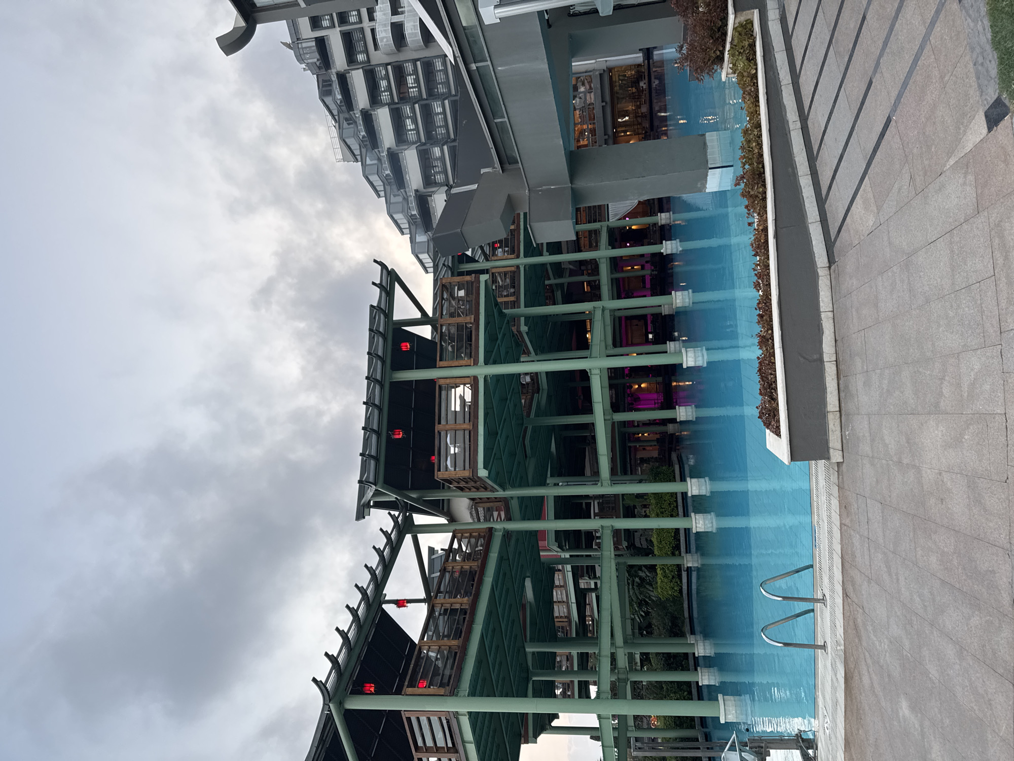 The image shows a large outdoor swimming pool with clear turquoise water. Metal railings lead into the pool from the tiled walkway. On the left, a multi-level structure with a dark roof and green supports is visible, with red lanterns hanging from it. In the background, a modern, stepped building with many windows rises into the cloudy sky. The scene appears to be a resort or hotel in Aksu, Turkey. The overall lighting suggests it is late afternoon or early evening. There are no people visible in the image.