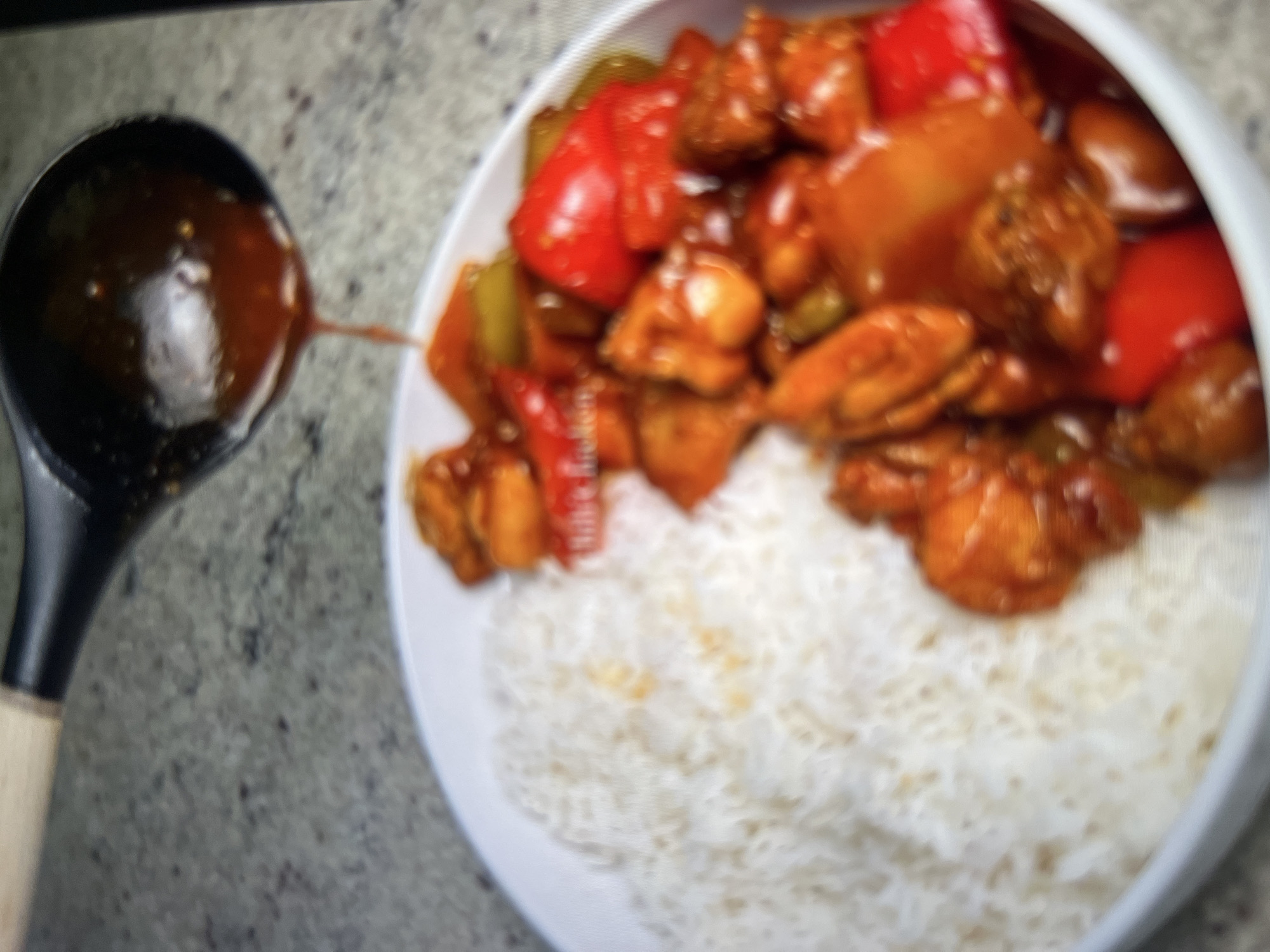 A ladle is pouring a dark, reddish-brown sauce over a white bowl filled with fluffy white rice and a stir-fry of chicken, red bell peppers, and green bell peppers. The scene is set on a speckled grey countertop. The image is blurry and lacks detail, making it impossible to determine the specific location in Abuja, Nigeria. There are no people visible in the image, and no specific actions or interactions are taking place beyond the pouring of the sauce. The environment is limited to the countertop and the bowl of food.