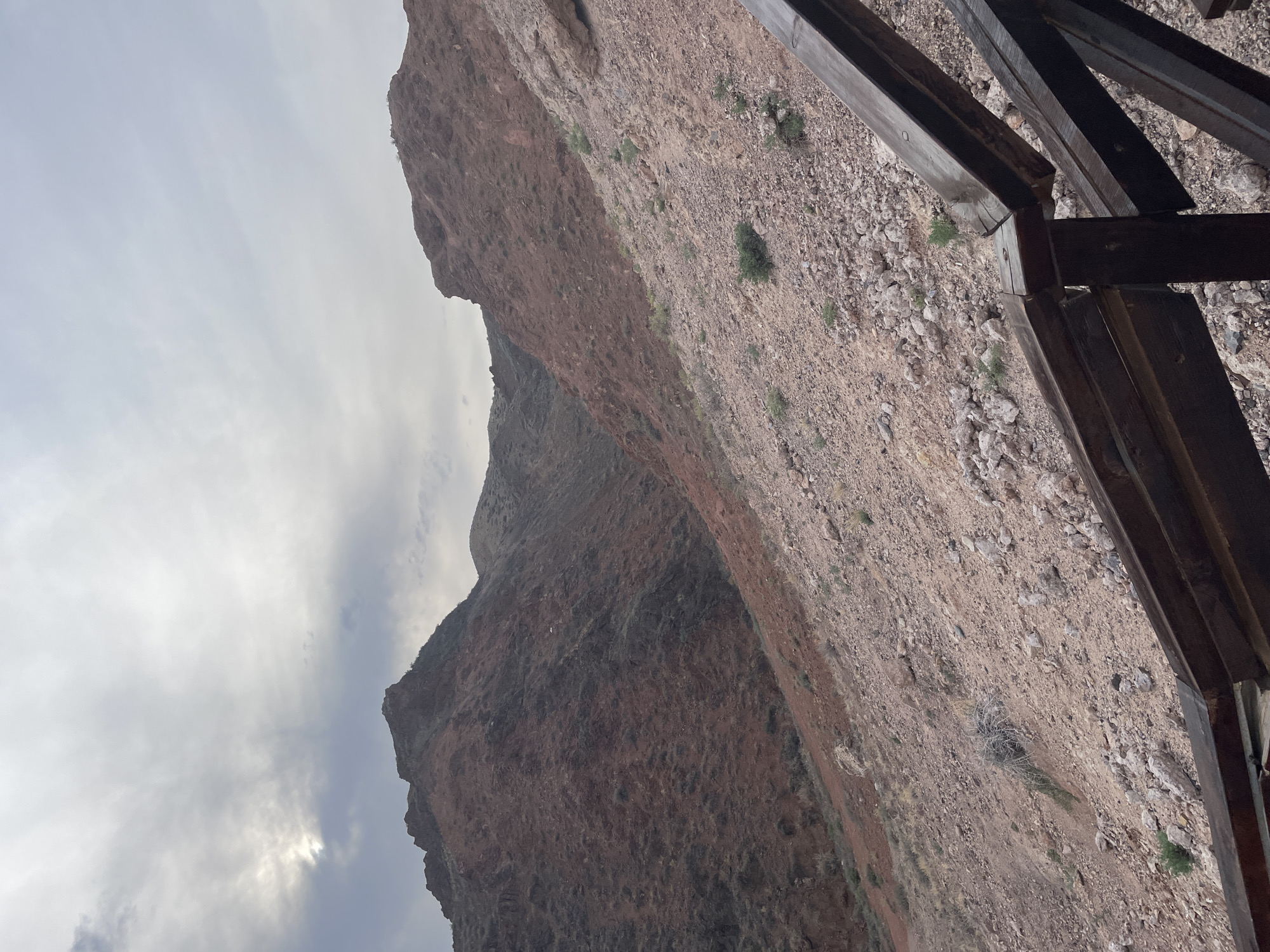 The image depicts a view of a mountainous, rocky terrain under a cloudy sky. The mountains are a reddish-brown color, with sparse vegetation. In the foreground, a portion of a wooden railing is visible, suggesting a viewpoint or walkway. The environment appears arid and rugged. The provided location "Tamerlik, Kazakhstan" does not appear to be a recognized city or significant geographical area in Kazakhstan based on available information.