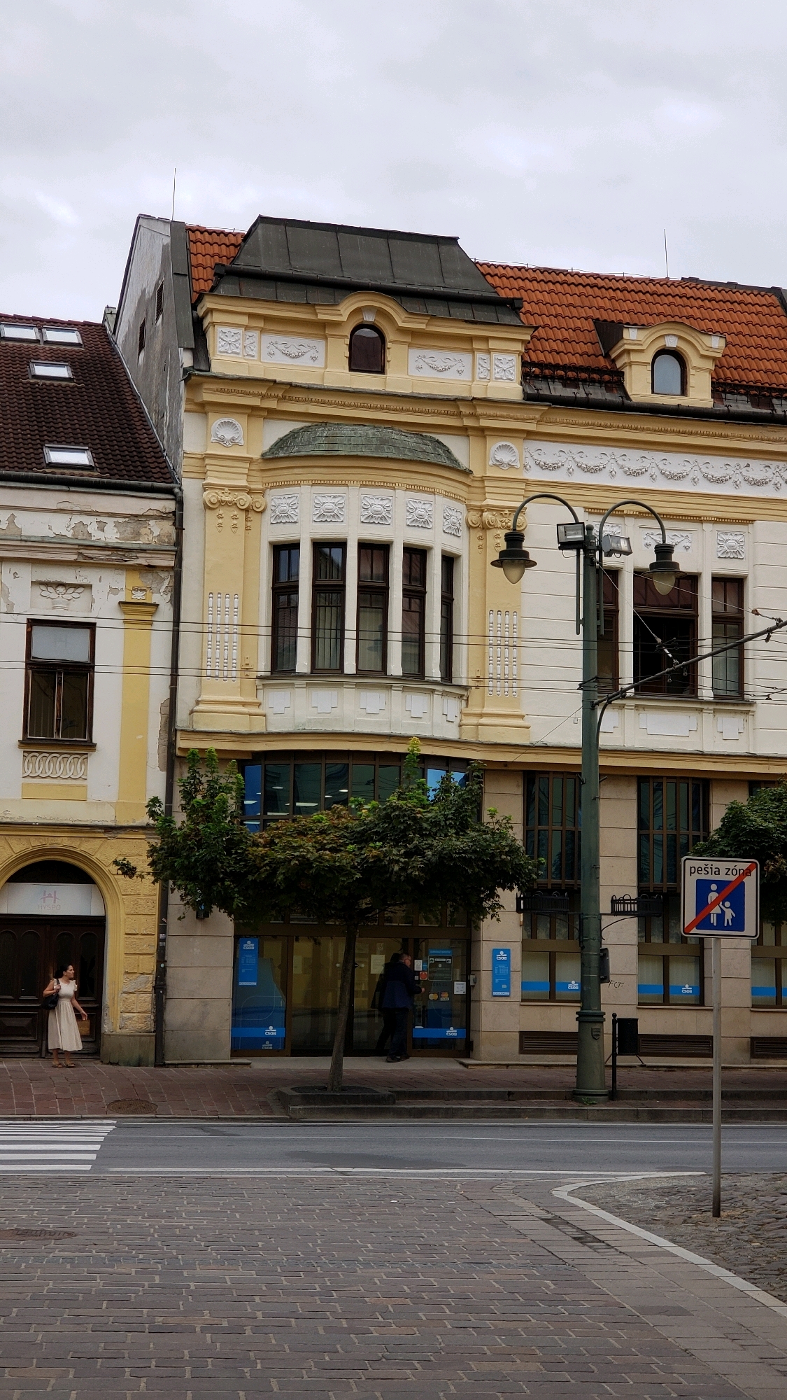 The image shows a street view in the city of Prešov, Slovakia. In the foreground, there is a cobblestone street with a pedestrian crossing. Behind the crossing is a sidewalk with a tree and a lamppost. To the right of the lamppost, there is a traffic sign that reads "pešia zóna" (pedestrian zone) with a symbol of a person and a child walking.
The buildings in the background are old, with ornate yellow facades and red tiled roofs. The building on the right has a bay window on the second floor and a modern glass storefront on the ground floor. A woman in a light-colored dress is standing on the sidewalk in front of a building entrance on the left. Two men are visible near the glass storefront of the building on the right, one entering and one exiting. The sky is overcast.