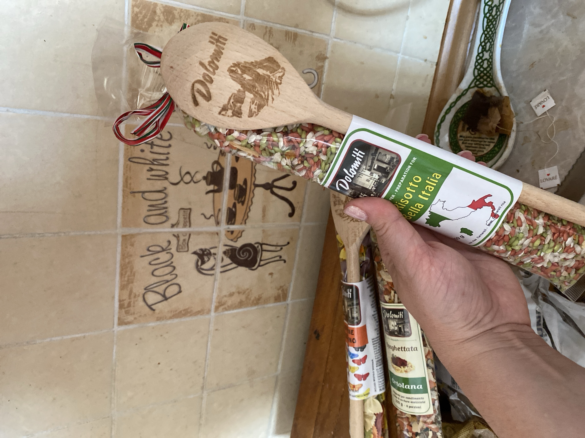 A hand is holding a wooden spoon with the brand name "Dolomiti" and an image of mountains carved into it. The spoon is inserted into a clear plastic bag filled with dry mixed grains and spices. The bag has a label that also reads "Dolomiti" and "Risotto Bella Italia." The background consists of beige tiles with a decorative mural featuring black and white drawings of cats, teacups, and a table. Several other similar products, also containing dry grains and spices, are visible beneath the spoon. Two small tea bags with "LOVARE" printed on them are on a ceramic spoon rest in the foreground. The image depicts a kitchen or pantry setting.