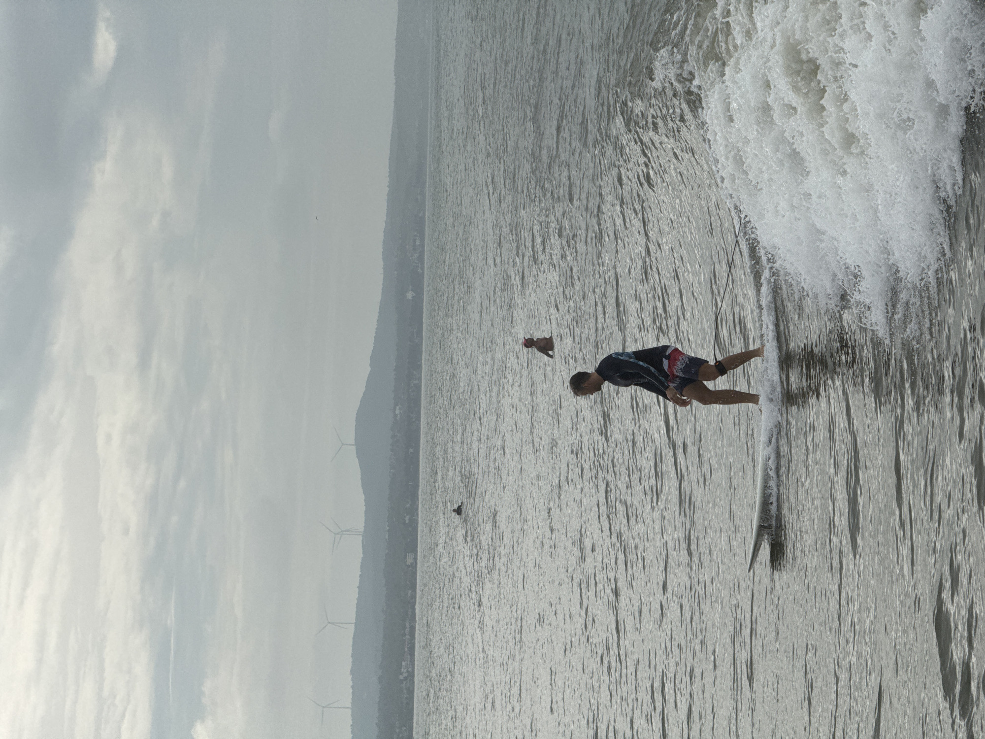 A medium shot captures a surfer in motion on a white surfboard in the ocean near Phan Thiết, Vietnam. The surfer, a male with fair skin, is wearing a dark, long-sleeved rash guard with blue and white striped accents on the sleeves and patterned board shorts. He is positioned in the center of the frame, riding a small wave. His body is turned slightly to the left, and his right leg is forward. To the right of the surfer, a frothy wave is breaking. In the distance, two other individuals are in the water, one appearing to be sitting on a surfboard and the other floating. The ocean is calm with gentle ripples, and the sky is overcast with heavy clouds. In the background, a hazy, mountainous coastline is visible, with several wind turbines dotting the ridge. Buildings are scattered along the coast. The overall lighting suggests it is daytime, likely late afternoon or early morning due to the soft, diffused light.