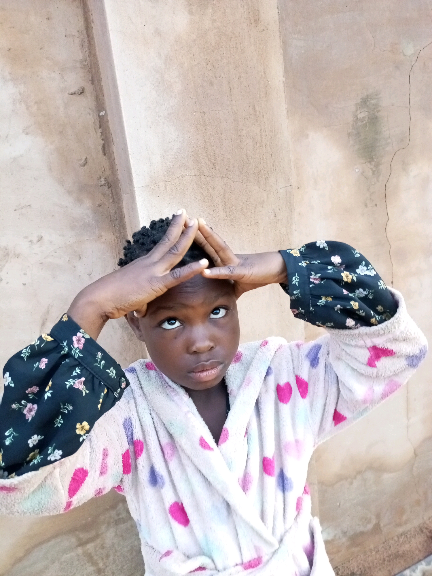 A young girl, appearing to be around 5-7 years old, is positioned in the foreground against a textured, light brown wall. She has dark skin and dark, curly hair, styled short. Her eyes are wide open, looking upward and slightly to her right. Her hands are clasped together above her head, forming a triangular shape with her fingers intertwined. She is wearing a light-colored bathrobe adorned with colorful heart-shaped patterns in pink and purple. The sleeves of the bathrobe are black with a floral print of pink and yellow flowers. The background wall shows signs of wear, with a prominent vertical seam or joint and a few small cracks. The lighting appears to be natural, casting subtle shadows. The overall impression is a candid portrait, likely taken outdoors due to the natural light and textured wall. The scene is a simple outdoor setting in what could be a courtyard or alleyway. The location is identified as Tempure, Nigeria.