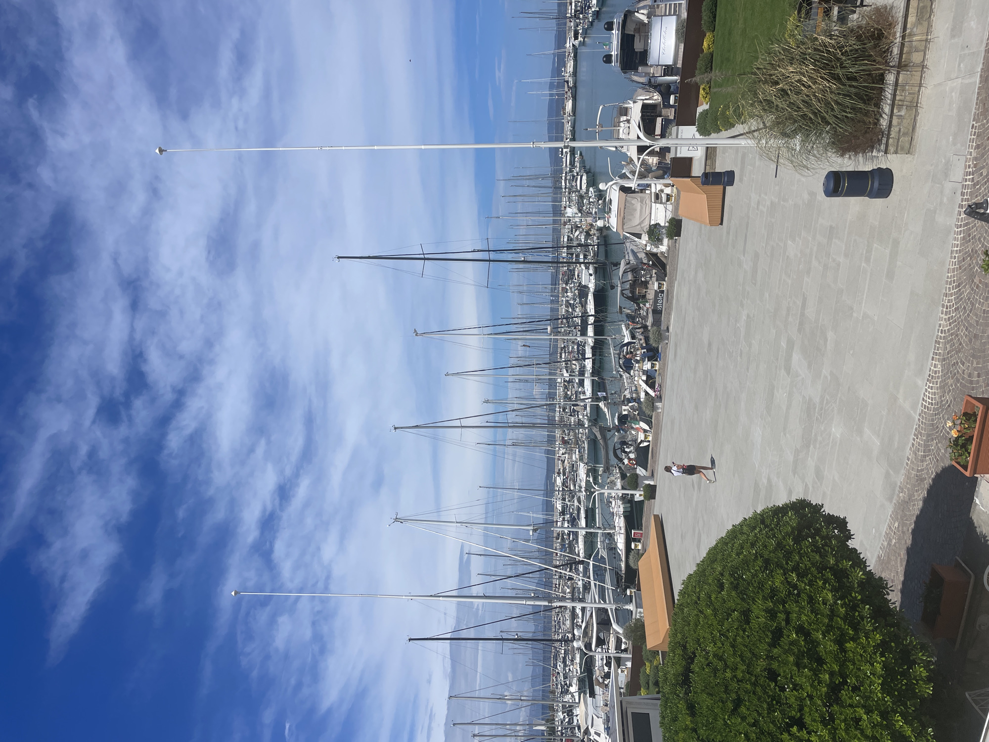 The image depicts a bright, sunny day at the Punta Ala marina in Italy, characterized by a vast blue sky with scattered white clouds. A multitude of sailboats and motor yachts are moored in the harbor, their masts forming a dense forest against the horizon. In the foreground, a paved plaza with stone tiles is visible. A single woman in a white top and dark shorts is walking across the plaza. Adjacent to the plaza are landscaped areas featuring lush green bushes and manicured lawns, as well as wooden benches and dark-colored trash receptacles. The scene conveys a sense of tranquility and leisure typical of a Mediterranean seaside resort.