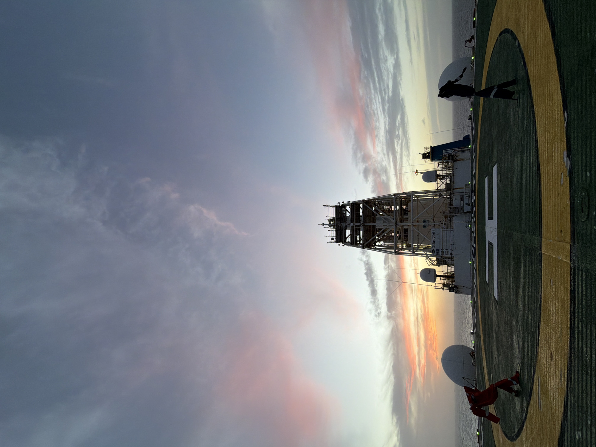The image shows an oil drilling ship, possibly the Globetrotter II, at sunset. The ship features a tall drilling derrick and a helipad marked with a white "H" inside a yellow circle on a green surface. Two satellite domes are visible, one near a person in red clothing standing on the helipad's edge. The sky displays varied cloud cover with colors ranging from purple and grey to orange and yellow, reflecting the sunset. The surrounding environment is the ocean, with a portion visible along the ship's edge. The location and city are unknown.