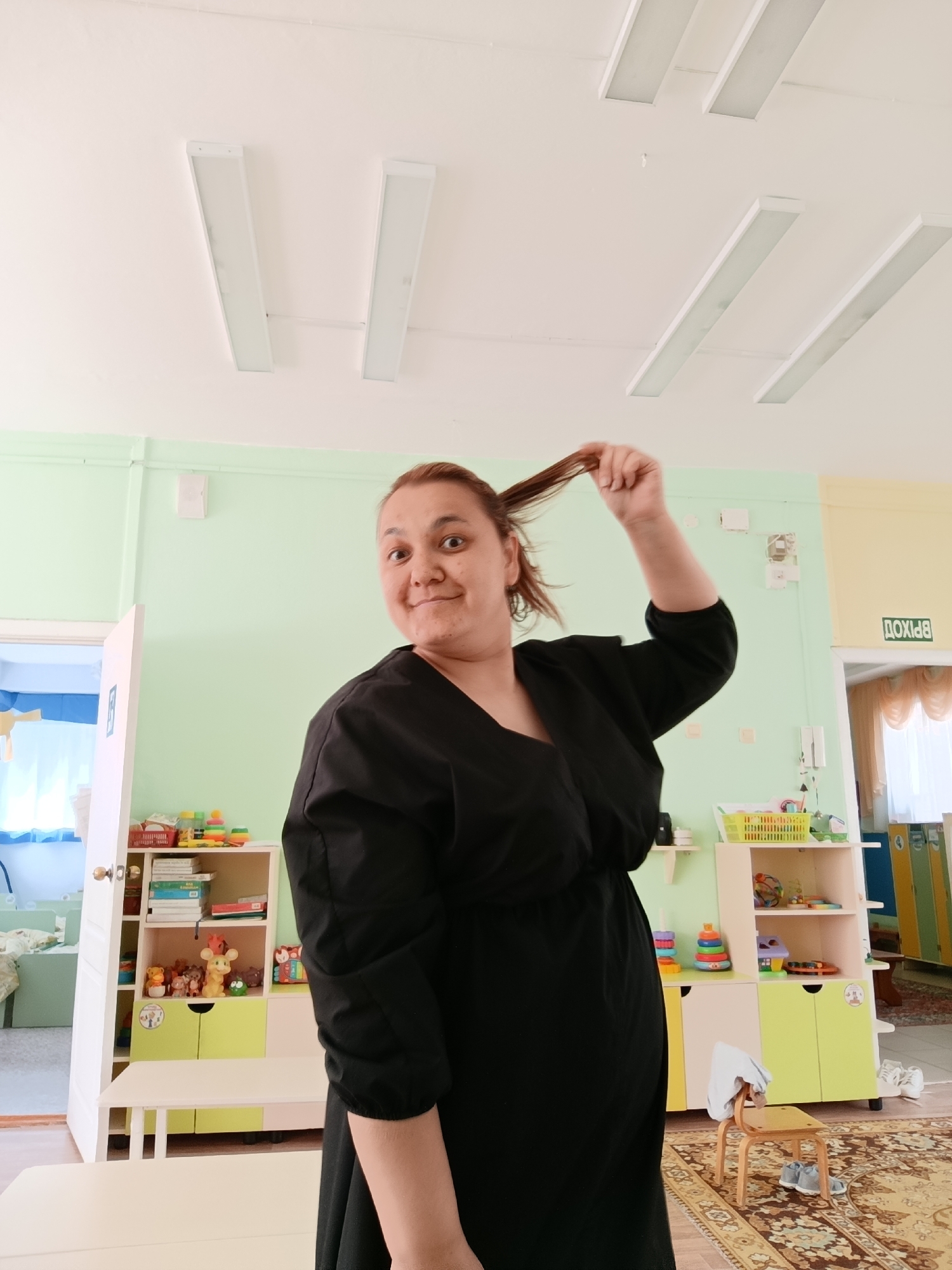 The image shows a woman in a black dress standing in a room that appears to be a classroom, possibly in a kindergarten or daycare setting. She is holding a section of her hair up with her right hand and looking at the camera with a slight smile. The room has light green walls, a white ceiling with long, rectangular light fixtures, and colorful furniture and toys. A door with a sign that reads "ВЫХОД" (EXIT) in Russian can be seen in the background. Based on the signage in Russian, the location may be in Chaikovsky, Russia.
