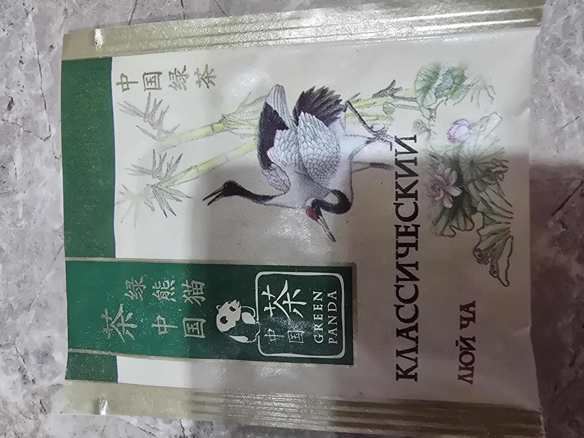 This image is a close-up, top-down view of a tea package. The package is mostly white with a green band across the top. On the left side of the package, there are vertical green bands with Chinese characters. Below these, there is an illustration of a panda sitting on a square. Inside the square are more Chinese characters and the English words "GREEN PANDA". To the right of the panda illustration, there is an illustration of a crane standing among bamboo stalks. Across the bottom of the package, there are Russian words: "КЛАССИЧЕСКИЙ" and "ЛЮЙ ЧА". The background of the image is a textured, grey surface that appears to be a countertop or table. There is no indication of the city of Ulan-Ude, Russia, in the image.