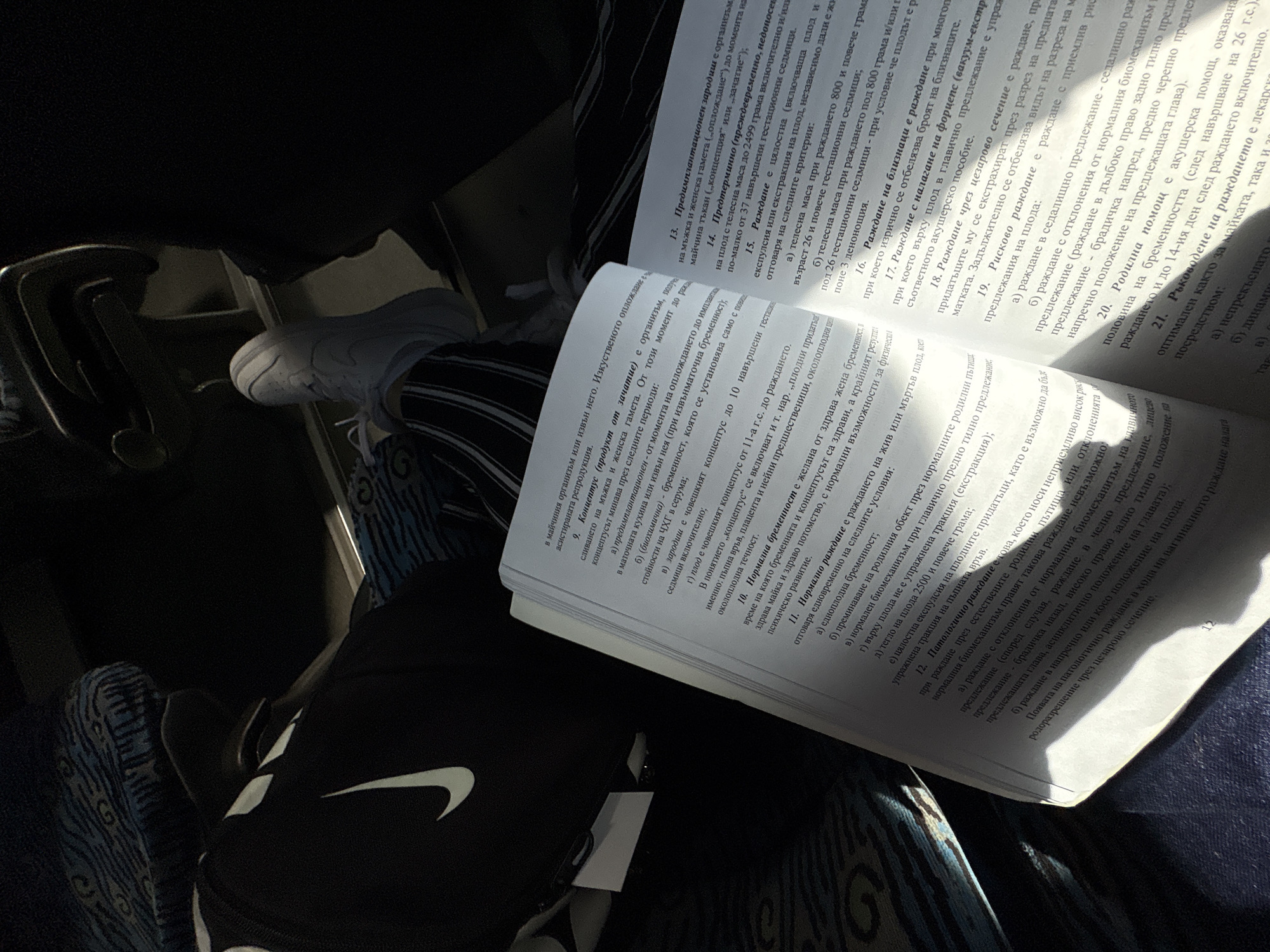 A person is sitting, presumably on a train or bus, reading a book. The person is wearing dark pants with white vertical stripes and white sneakers. The book is open and lying on their lap. The text in the book is in Bulgarian. The book appears to be a medical text, likely concerning childbirth or gynecology. There is a black Nike bag near the left side of the image. The location appears to be indoors in transit, potentially in Byala Slatina, Bulgaria, but this is speculative based on the Bulgarian language in the book.