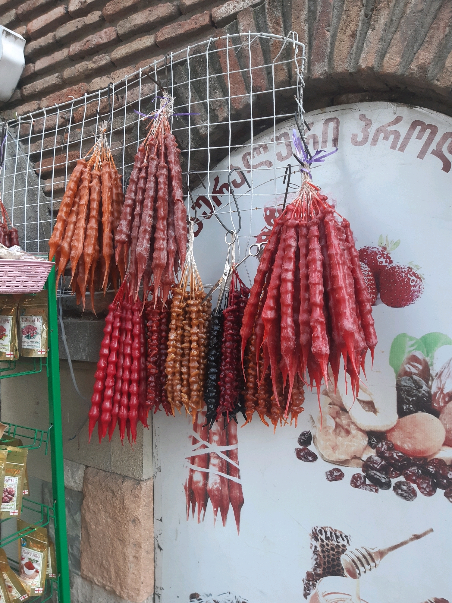 The image depicts a close-up view of a street vendor's stall in Tbilisi, Georgia, displaying a variety of traditional Georgian churchkhela candies. These candies, made from grape must and nuts, are strung together and hung from a wire grid. They are arranged in numerous bunches, showcasing a range of colors from deep red to reddish-brown and lighter brown. In the background, a textured brick wall and a white surface with colorful illustrations of fruits, nuts, and a honeycomb are visible, suggesting an outdoor market or a shop facade. Packaged goods in golden wrappers are also displayed on a green metal rack to the left. The overall scene is bright and colorful, highlighting the artisanal nature of the products.