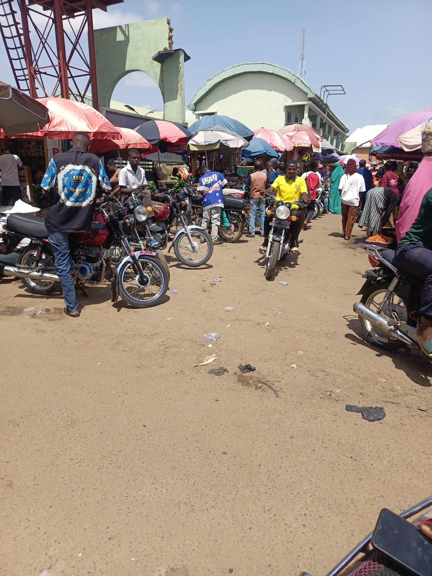 The image shows a bustling street scene in Akwanga, Nigeria. Several motorcycles are present, some stationary and some with riders navigating through the crowd. The people are dressed in various casual attires. Parasol umbrellas are scattered throughout, likely providing shade for vendors and customers. The road surface is paved but shows signs of wear and scattered debris. The architecture in the background includes buildings with arched entrances and a water tower, suggesting this is a busy market area.