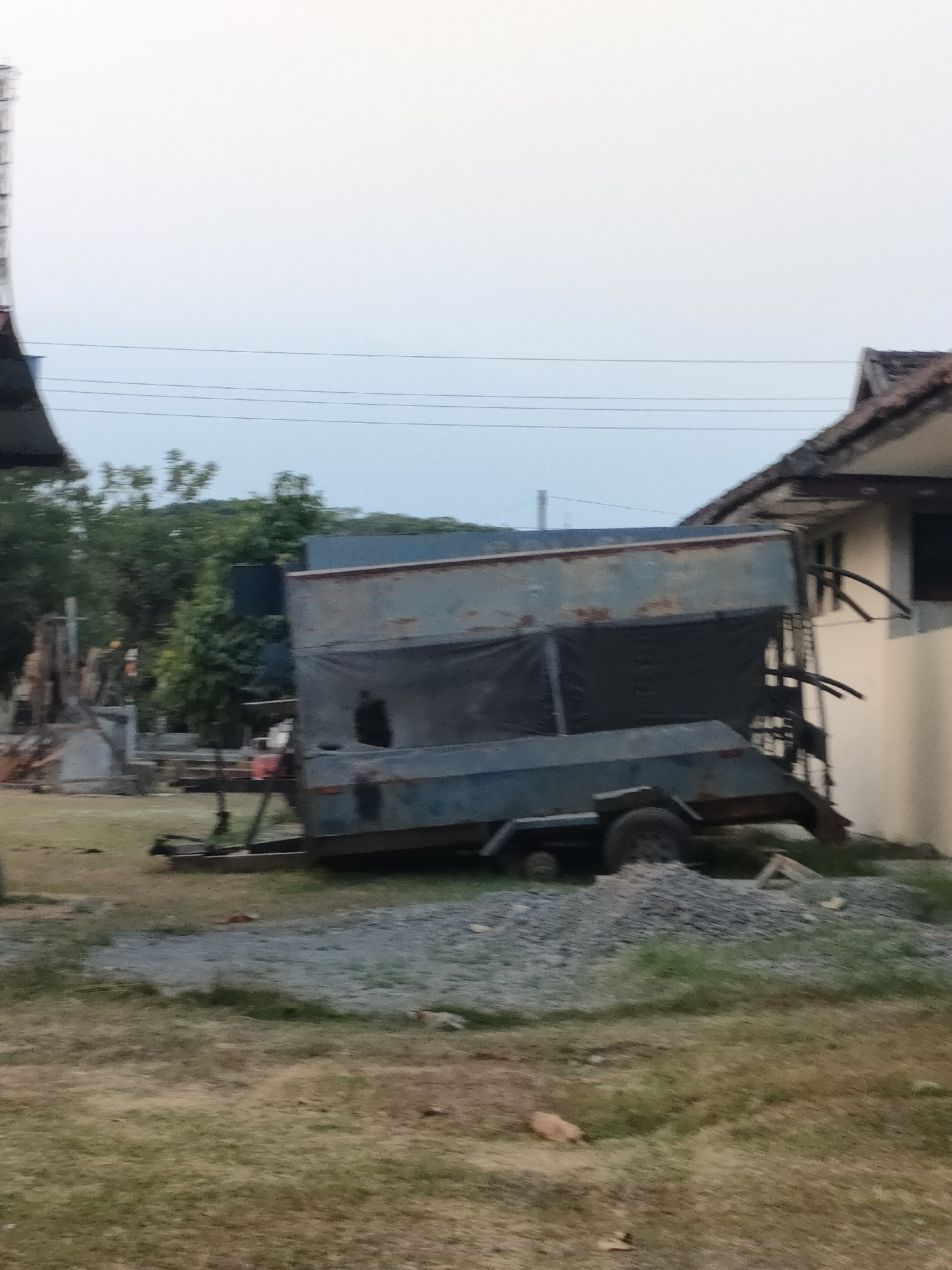 In Banguntapan, Indonesia, a trailer with a large, rusted metal enclosure covered by black tarp is parked on a grassy area next to a building. The trailer has two wheels visible on the left side and a ramp at the rear. The building appears to be made of concrete with a tiled roof and has at least one window visible. In the background, there are trees and utility lines. The ground is covered in grass and gravel.