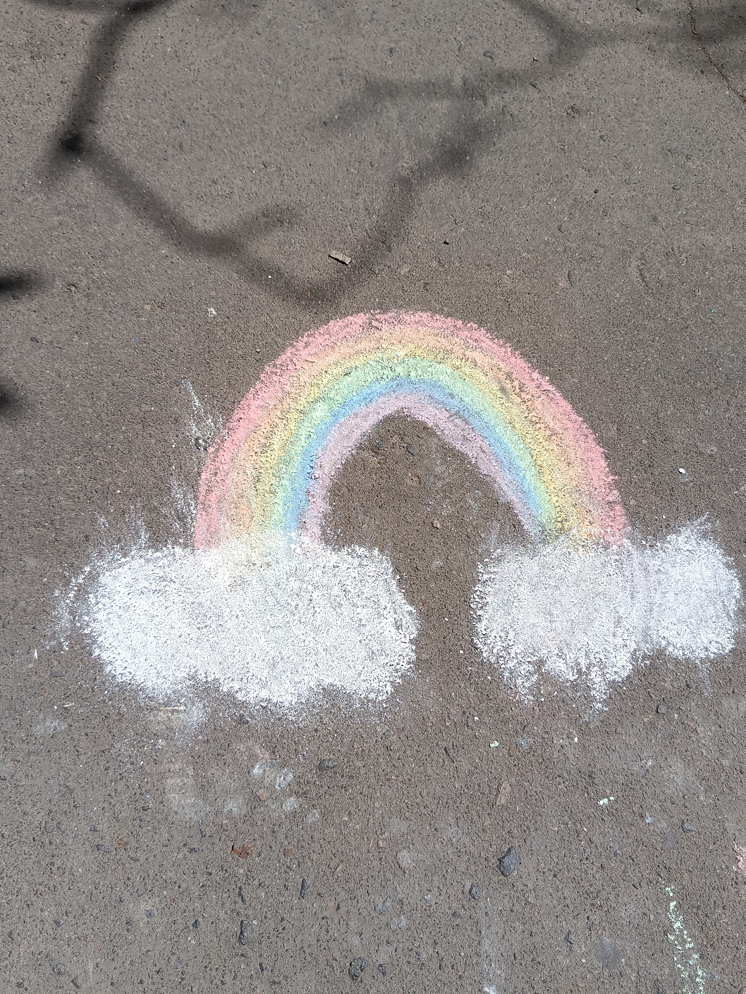 A chalk drawing of a rainbow with clouds is on an asphalt surface. The rainbow arcs from the left side of the image to the right, with bands of pink, yellow, and blue chalk. White chalk forms two cloud shapes at the base of the rainbow. Shadows of tree branches are visible on the asphalt in the upper portion of the image.