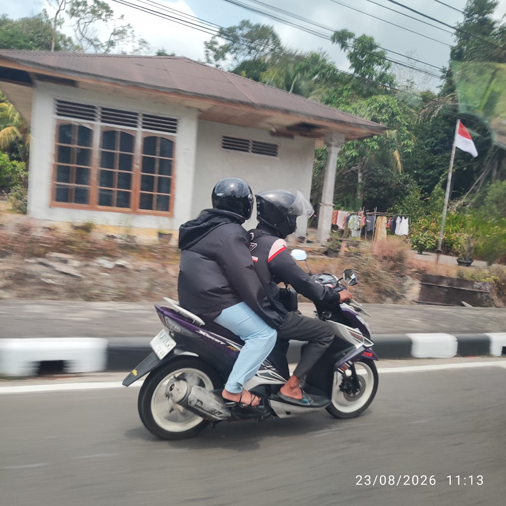 Two individuals wearing black helmets are riding a dark purple and white motorcycle on a paved road. The person in the front is wearing a black jacket and dark pants, while the person in the back is wearing a black hooded jacket and light blue pants, and black sandals. They are both facing forward. The motorcycle has a license plate with the inscription "BN 6129 JS 12.26". In the background, there is a building with a brown roof and windows, surrounded by trees and vegetation. A red and white flag is visible on a pole to the right of the building. The scene appears to be in a rural or suburban area. The timestamp on the image reads "23/08/2026 11:13". The location is identified as Lesung Batang, Indonesia.