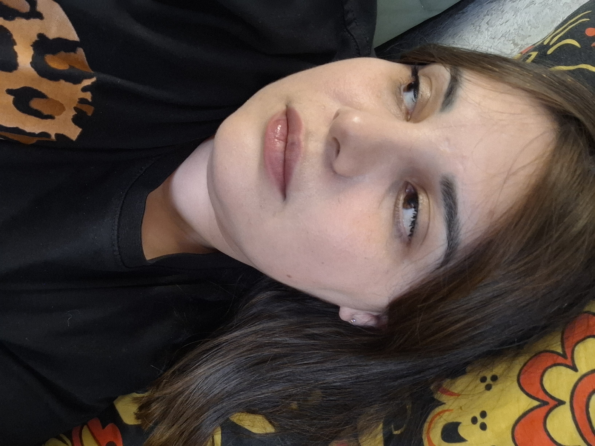 A close-up, tilted portrait shows a young woman lying down. Her head rests on a patterned surface, possibly a pillow or blanket, which features a vibrant yellow background adorned with red-orange and black floral or abstract designs. Her dark brown hair is spread out beneath her head and flows along her right shoulder.
The woman has fair skin, brown eyes, and dark eyebrows. She wears subtle makeup, including a light, shimmery eyeshadow and glossy, pale pink lipstick. Her expression is serene and neutral, with her gaze directed slightly upwards and to her right. A small, silver stud earring is visible in her right earlobe.
She is dressed in a black top or t-shirt, which features a large, partially visible leopard print pattern in brown and black on the upper left side. The indoor setting, suggested by the patterned fabric and relaxed pose, appears to be a comfortable space, possibly a bedroom or living area. No text is visible in the image, and details such as time of day or weather are not discernible.