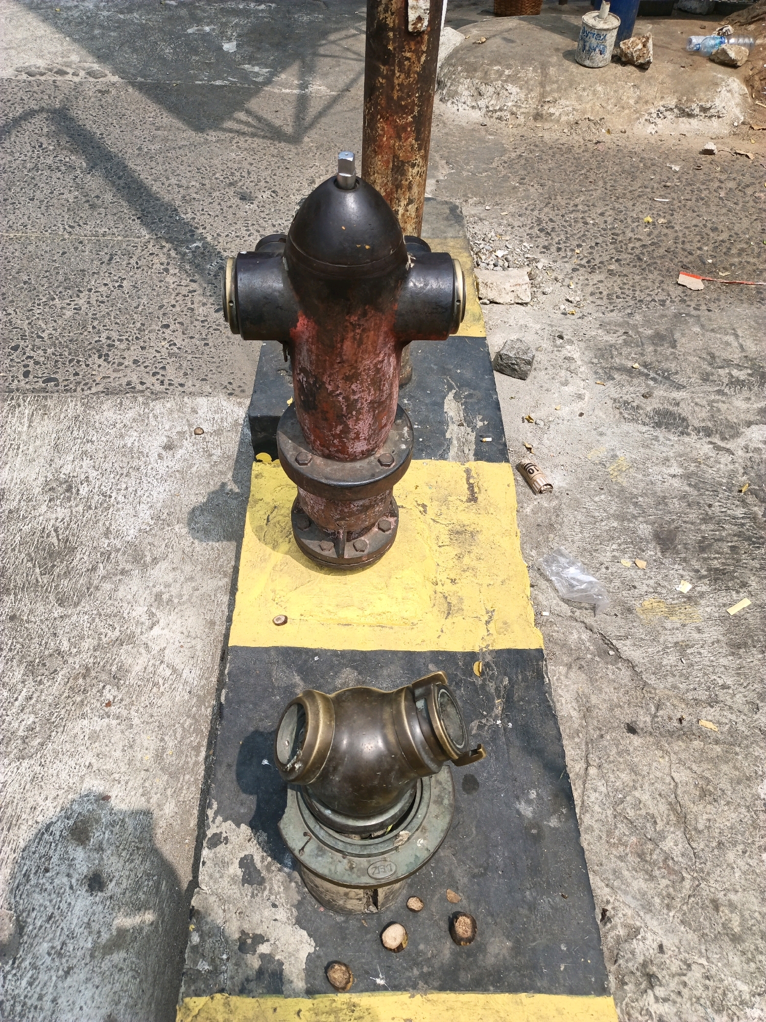 The image shows a fire hydrant painted red and black, standing on a yellow and black striped platform. In front of the hydrant, a separate brass fitting, possibly a cap or connector, rests on a round metal base. The scene appears to be outdoors on a paved surface, with a road and some debris visible in the background. The area suggests an urban environment.