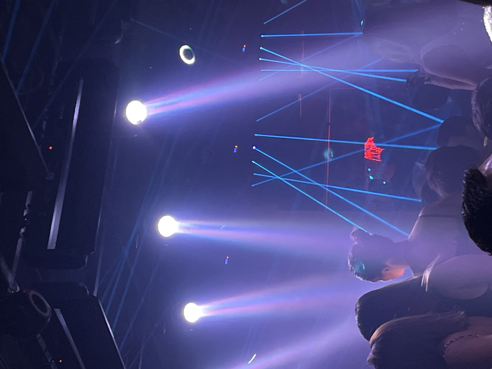 The image depicts an event at a location appearing to be a nightclub or concert venue in San Bernardino Tlaxcalancingo, Mexico. Several bright spotlights shine beams of light into the crowd and across the room, creating a foggy, atmospheric effect. Blue laser beams cut through the air. Silhouettes of people are visible in the foreground, particularly on the right, suggesting a dense crowd. A red object is suspended or projected in the background. The overall environment appears dimly lit with a focus on the stage and light effects. Structural elements like metal scaffolding are visible on the left.