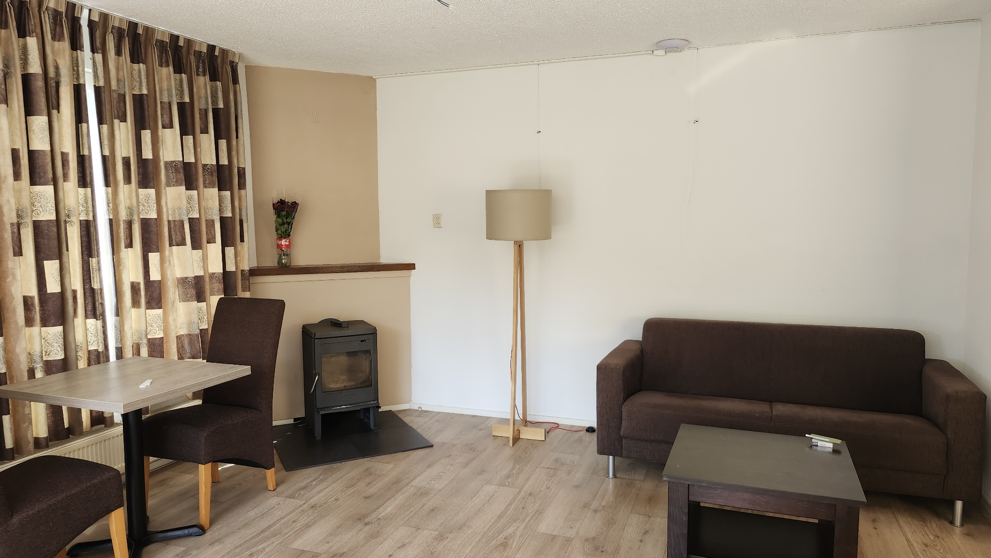This is a wide-angle, eye-level, interior shot of a living room in Exloo, Netherlands. The room features light-colored wood-plank flooring and white walls, with a beige accent wall on the left. In the foreground, a light brown square table with a black pedestal base is positioned with a matching chair and a bench-like seat behind it, all upholstered in dark brown fabric. To the right of the table is a black wood-burning stove on a dark gray hearth. Behind the stove, a shelf is visible with a vase of red roses and a red Coca-Cola bottle. Further to the right, a floor lamp with a beige cylindrical shade and a light wood tripod base stands near a dark brown two-seater sofa. A dark brown coffee table is situated in front of the sofa, with a pack of cigarettes and a lighter on its surface. Curtains with a geometric pattern of brown and beige squares hang on the left side of the room. A white ceiling with visible light fixtures and a smoke detector is present.