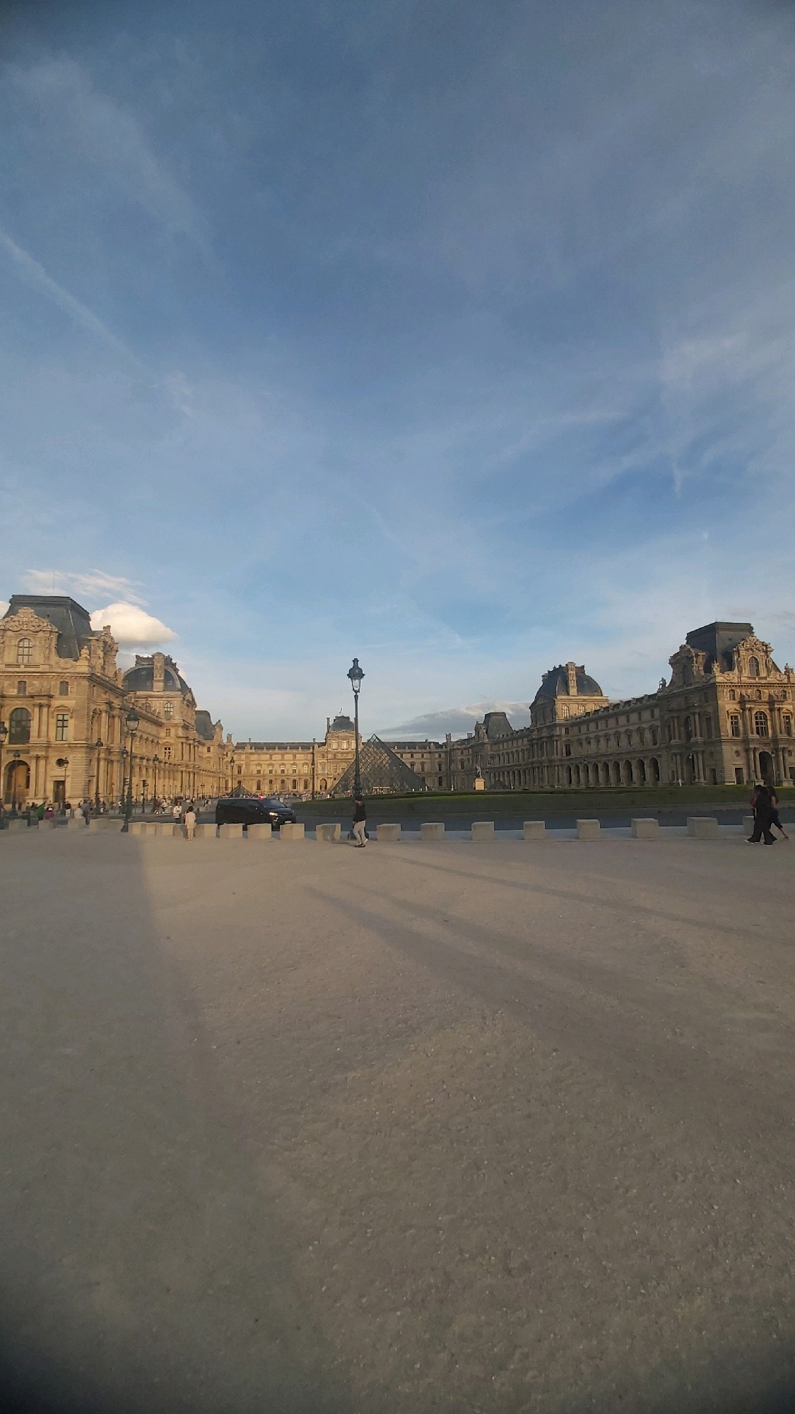 The image depicts the Louvre Museum in Paris, France, under a partly cloudy sky. The vast courtyard in front of the museum is paved with gravel and features long shadows cast by unseen objects or structures. A black van is parked in the middle ground. Several people are visible in the scene; some are walking across the courtyard, and others are gathered near the museum's facade. A prominent lamppost stands in the foreground, and the iconic glass pyramid of the Louvre is visible in the distance. The architecture of the museum, characterized by its classical style with ornate details, dominates the background. The overall lighting suggests it is late afternoon or early evening, as indicated by the long shadows.