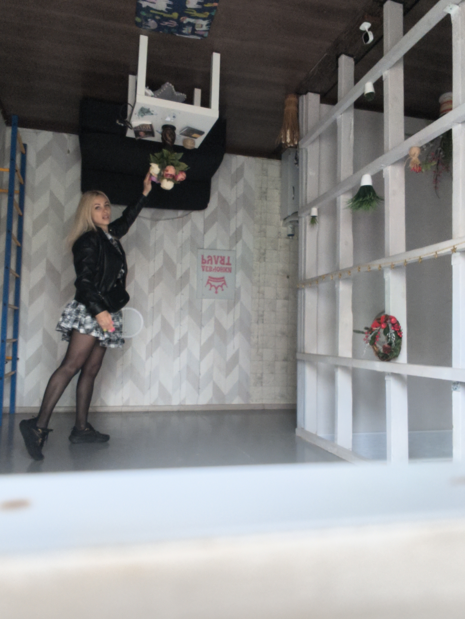 This image depicts a blonde woman posing inside an "upside-down house" illusion room, a type of interactive exhibit common in places like Sevastopol, Ukraine. The room is designed to create an optical inversion, with the floor appearing as the ceiling and furniture fixed overhead.
The woman is centered in the frame, standing on a wall with a distinctive gray and white chevron pattern. She wears a black leather jacket over a plaid skirt, black tights, and black sneakers. Her right arm is extended upwards, holding a bouquet of pink and white flowers towards an inverted white table and a black sofa, which are positioned on the "ceiling" of the room. A white chair is also attached to the ceiling near the table.
To the right, a large white shelving unit or partition is filled with various decorative elements, including a red and green wreath, small green plant ornaments, and other small items. On the far left, a blue and yellow ladder or set of bars is visible. A pink sign is affixed to the chevron wall, displaying mirrored text: the upper word reads "LAVRAT" (the reverse of "TRAVEL"), and below it, another word appears in mirrored Cyrillic script, accompanied by a crown symbol.
The indoor setting is evenly lit, and the overall scene conveys a sense of playfulness and visual disorientation, typical of an optical illusion exhibit.