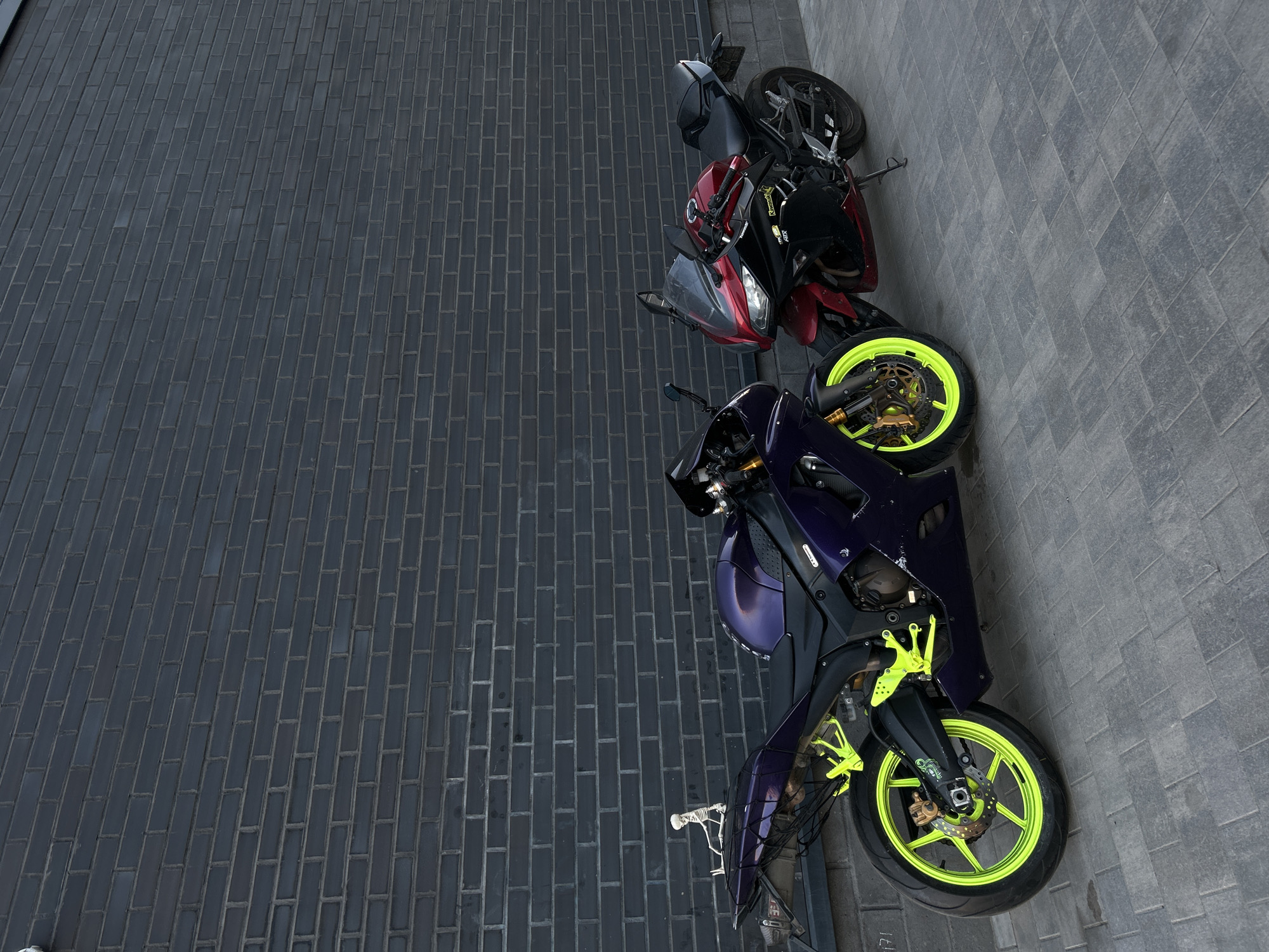 The image depicts two motorcycles parked on a gray brick surface. One motorcycle is dark purple with bright green rims, while the other is primarily red and black. The brick surface appears to be part of a street or parking area. Based on the architectural style and the presence of certain vehicle types, it's possible the location is in Saint Petersburg, Russia. No people are visible in the image.