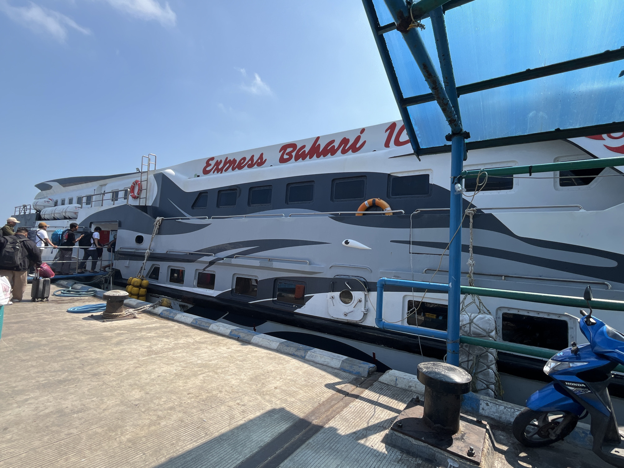 An express boat named "Express Bahari 10" is docked at a pier in Karimunjawa, Indonesia, under a clear blue sky. Several passengers with luggage are waiting on the pier to board. The boat is white with grey accents and red lettering. A blue and green canopy covers part of the pier. A blue scooter is parked near the boat. The pier is made of concrete with a textured surface and a blue and white border. A thick rope is secured to a bollard on the pier, connecting to the boat. Yellow buoys are attached to the side of the boat.