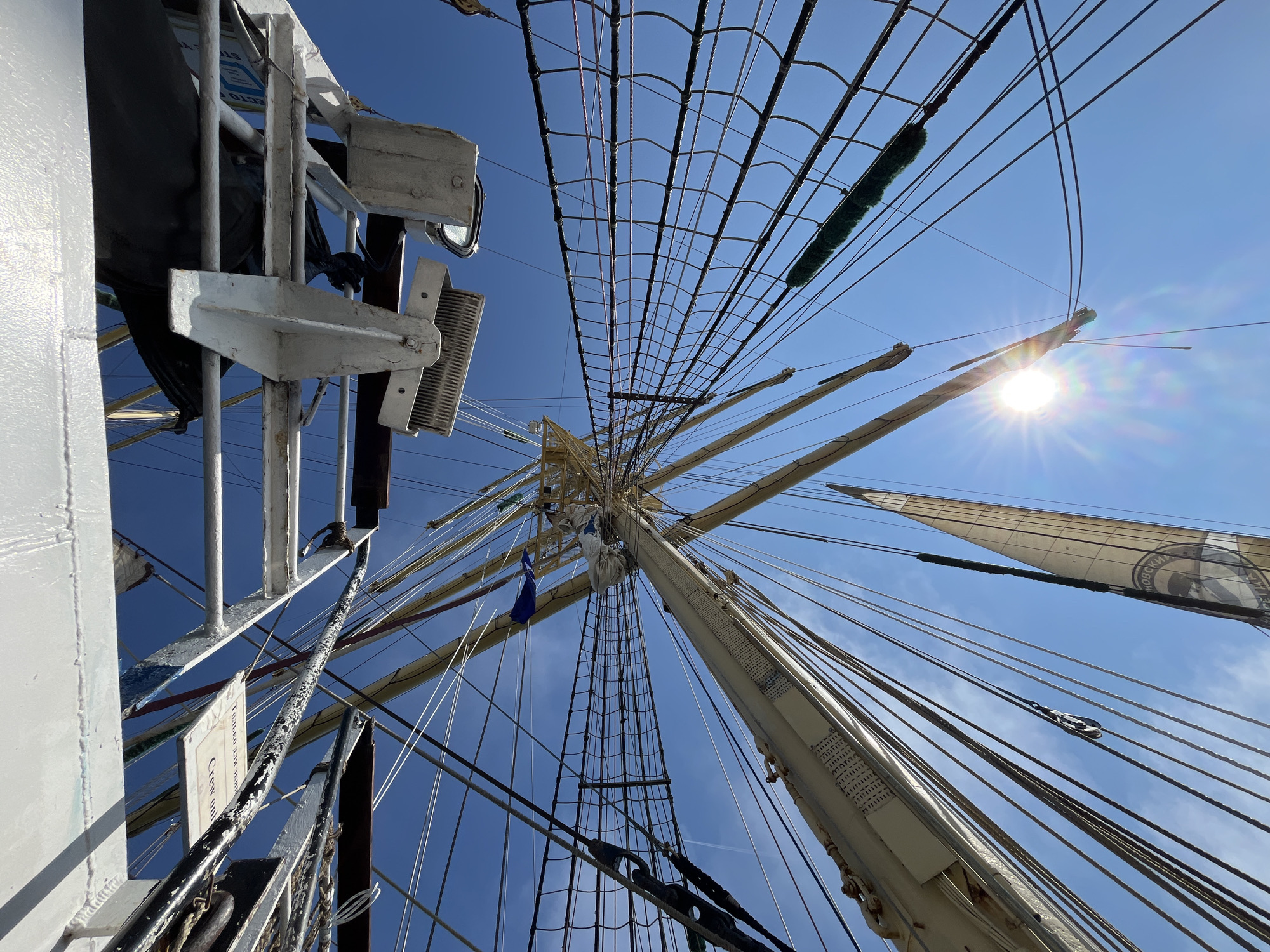 The image shows a low-angle view of the rigging and masts of a tall ship against a clear blue sky. The sun is visible, casting a lens flare. Several sails are furled or partially deployed, and a dense network of ropes and lines crisscrosses the frame. Some metal structures and equipment are also visible, attached to the ship's superstructure. There are no people or animals visible in the image. The scene appears to be a sunny day at sea or in port. The location is identified as Pionersky, Russia, suggesting the ship is in or near this coastal city.