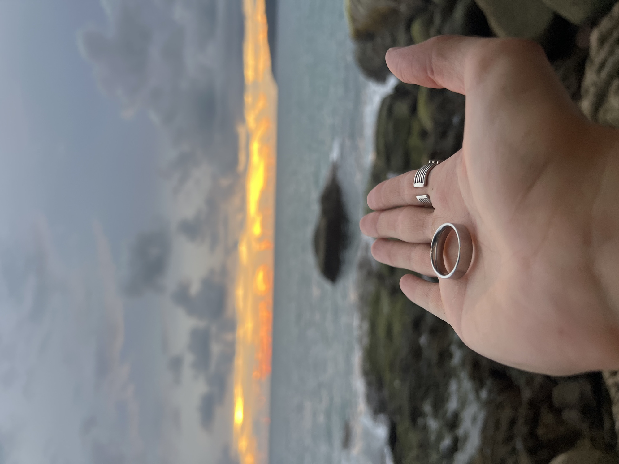 A close-up of an open palm facing the camera, displaying two silver rings. One ring is plain and smooth, resting in the center of the palm. The other, a decorative band with horizontal ridging, is worn on the ring finger. The hand is positioned in the foreground, with the middle ground featuring a rocky shoreline and the ocean. In the background, the sea meets the horizon, which is illuminated by a dramatic sunset casting yellow and orange hues across the sky. The sky is filled with dark, dramatic clouds. The location is Karon, Thailand.