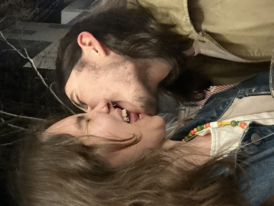 A couple, a man and a woman, are captured in a close-up shot appearing to kiss. The man has long dark hair and facial hair, and wears a tan jacket. The woman, with her mouth open, sports a nose ring and braces, and her long brown hair frames her face. She is dressed in a denim jacket over a white top and a colorful beaded necklace. The background includes dark, indistinct elements that suggest an outdoor setting, possibly with bare branches.