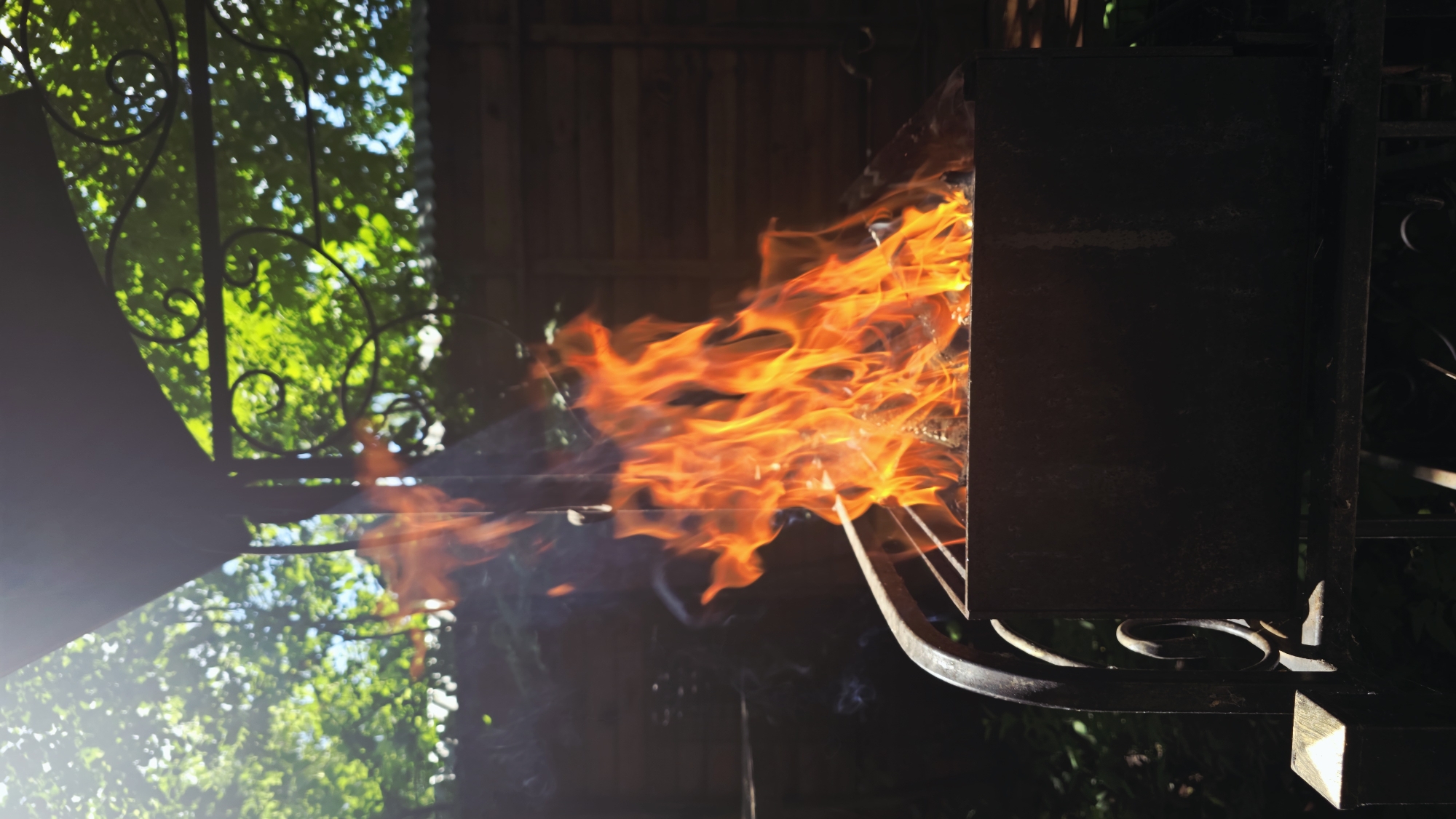 The image shows flames erupting from a metal barbecue grill, likely in a backyard setting. The fire is a vibrant orange and yellow, with tendrils of smoke rising from it. The grill is dark metal, and parts of it are obscured by the flames. In the background, there are green trees and a wooden fence, suggesting an outdoor location. The sunlight filters through the leaves, creating a dappled effect. There is also a decorative metal gate in the foreground. The overall impression is one of heat and intensity, with the natural elements of trees and light contrasting with the man-made fire. The time of day appears to be daytime, possibly late afternoon, given the angle of the sunlight. The location context indicates Il'inskiy, Russia.
