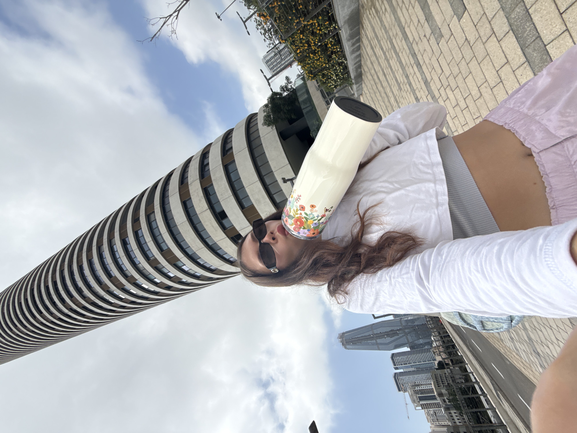 A woman wearing sunglasses and a white jacket is pictured holding a water bottle. The scene is from Tel Aviv, Israel, with the Azrieli Sarona Tower in the background.