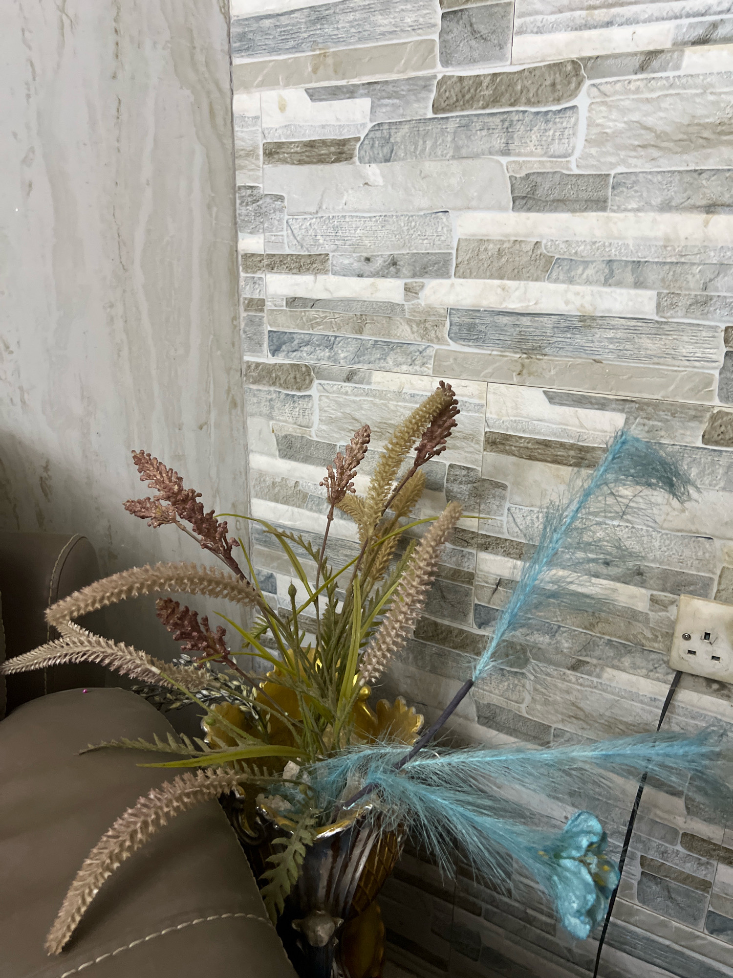 An interior scene depicts a faux floral arrangement in a decorative vase situated next to a plush brown sofa. The background features a wall with a stone-tile pattern in muted grey and white tones, and to the left, a wall with a marble-like texture in beige and white. A single electrical outlet with two sockets is visible on the patterned wall. The faux flowers are a mix of dried-looking stalks in light brown and reddish-brown hues, along with vibrant teal-colored feathery plumes. The overall impression is of a decorated living space. The location is identified as Dar es Salaam, Tanzania.