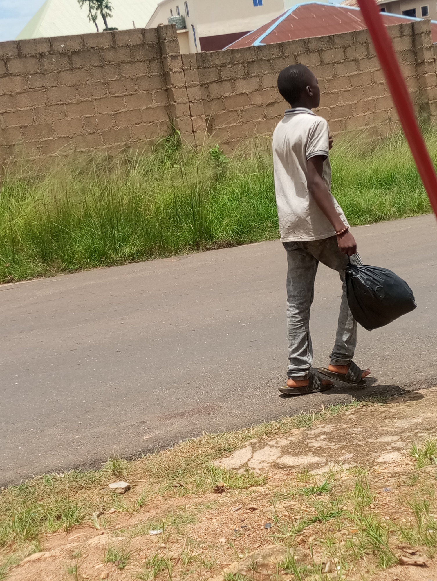 A young Black male walks along the side of an asphalt road, carrying a black plastic bag in his right hand. He is wearing a light-colored polo shirt and faded gray jeans, along with dark-colored sandals. His left arm hangs by his side. The background features a light-colored brick wall with a strip of tall green grass behind it. Beyond the wall, two buildings are visible; one has a red, sloped roof with light blue trim, and the other is a lighter-colored structure. A red pole is partially visible on the right side of the frame. The ground beside the road is rough, with patches of dry earth and sparse green grass. This scene is consistent with the environment found in Jos, Nigeria.