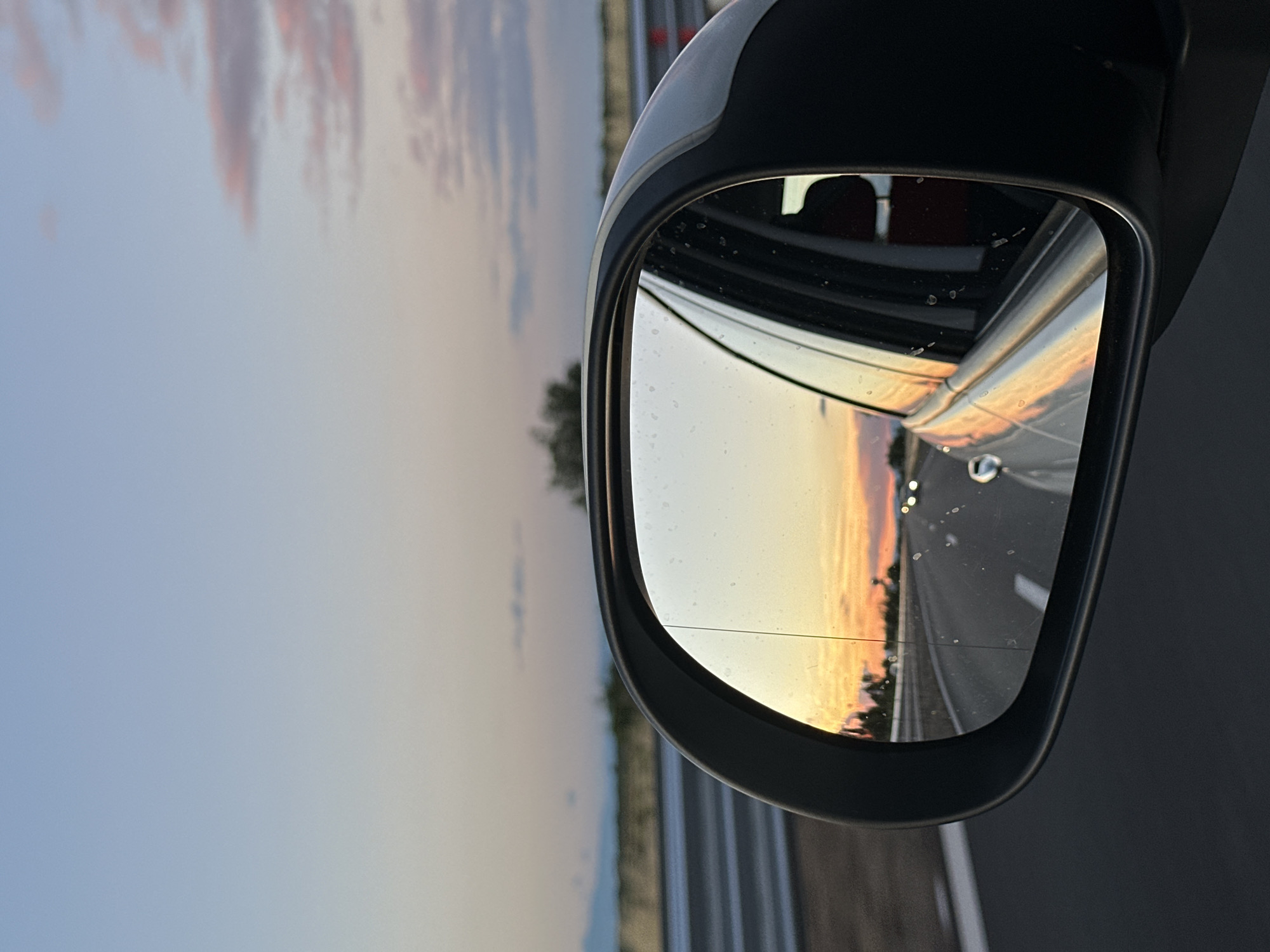 The image shows a side-view mirror of a car reflecting a sunset over a highway. The sky is a gradient of blue, pink, and orange. In the distance, there are faint outlines of mountains and trees. The highway is visible, with lane lines and other cars in the reflection. The car itself is white, and its interior is partially visible through the window. The image is taken from a high angle, looking down on the mirror. There are some water droplets on the mirror's surface. The location is identified as Malo Konare, Bulgaria.