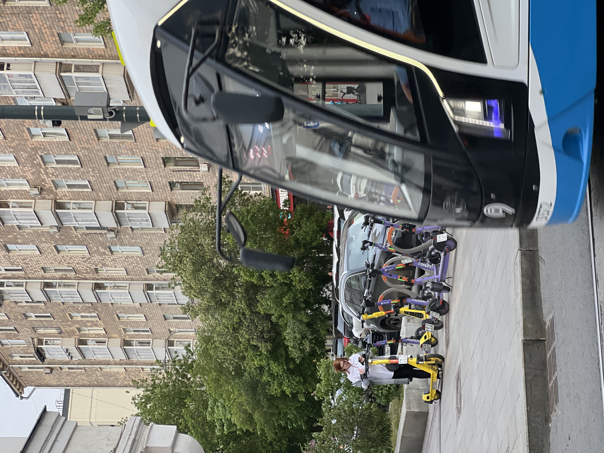 The image captures a street scene in an urban environment with a large building in the background featuring multiple windows. In the foreground, the front of a modern tram is prominently displayed. Near the tram and along the sidewalk, there are several parked electric scooters of varying colors, including yellow and purple. A person is standing near the scooters, holding one of the yellow ones, seemingly interacting with it. Additionally, a parked car is visible behind the scooters.