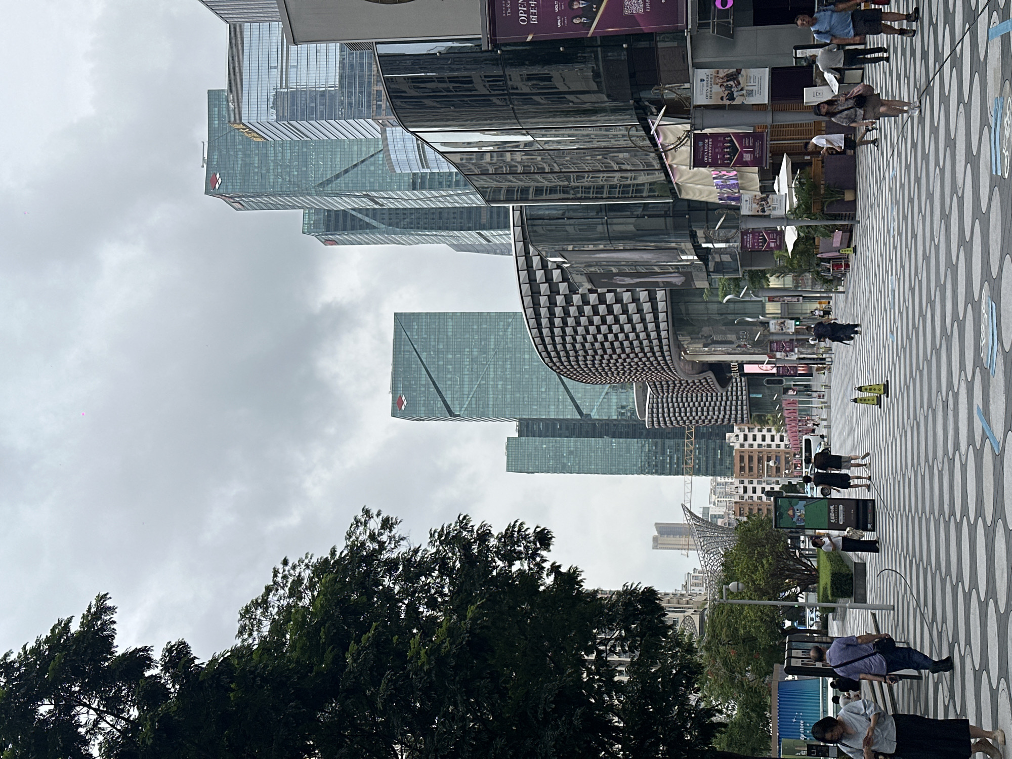 The image depicts a bustling street scene in Futian District, China, under a cloudy sky. The foreground features a wide, pedestrian-friendly walkway with a distinctive circular pattern on the paving stones. Several individuals are walking along the path, engaged in various activities such as strolling, carrying bags, and looking at their phones.
To the right, modern architecture dominates the scene, with a glass-fronted building featuring reflective surfaces and a unique, curved facade adorned with a checkered pattern. Banners advertising an "Open House" are hung from the building's exterior, suggesting commercial activity.
Further into the background, a cluster of tall, modern skyscrapers rises into the overcast sky, their glass exteriors reflecting the ambient light. The buildings vary in design, with some featuring sharp angles and others with more curved or faceted surfaces. Green trees line the left side of the street, providing a contrast to the urban landscape. In the distance, construction cranes are visible, indicating ongoing development in the area. The overall atmosphere is one of a vibrant, contemporary urban environment.