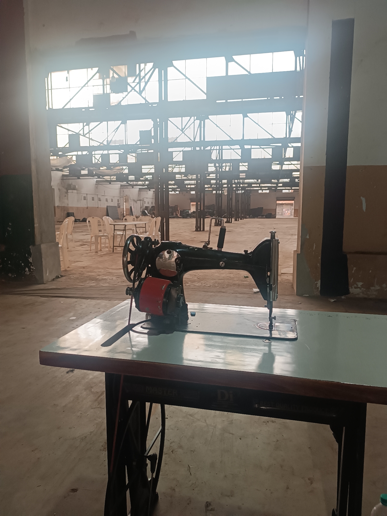 The image captures a large, empty industrial building or warehouse, with a prominent sewing machine in the foreground. The sewing machine is an industrial model, dark green and black, with a red motor casing, resting on a light green table. The table itself has a dark brown frame and features a large flywheel, characteristic of older industrial sewing machine stands. Text partially visible on the table frame reads "MASTER" and "DI", with fainter text below "DI" that seems to say "BEST QUALITY".
In the mid-ground, numerous white plastic chairs are arranged around a few tables, suggesting a communal area like a cafeteria or a meeting space, though it is currently unused. The floor throughout the vast building appears to be concrete.
The background reveals the immense scale of the building, with its high ceilings supported by metal structural beams and cross-bracing. Large windows or skylights across the far end of the structure let in abundant, bright daylight, creating a backlit effect that silhouettes much of the industrial framework and fills the upper portion of the image with a bright haze. This strong natural light indicates it is daytime. The walls of the building show signs of wear, with some areas appearing faded or with peeling paint. No people are visible in the scene, conveying a sense of quiet emptiness or a facility that is either disused, under renovation, or awaiting activity. The overall impression is one of a large, functional space currently in a state of repose.