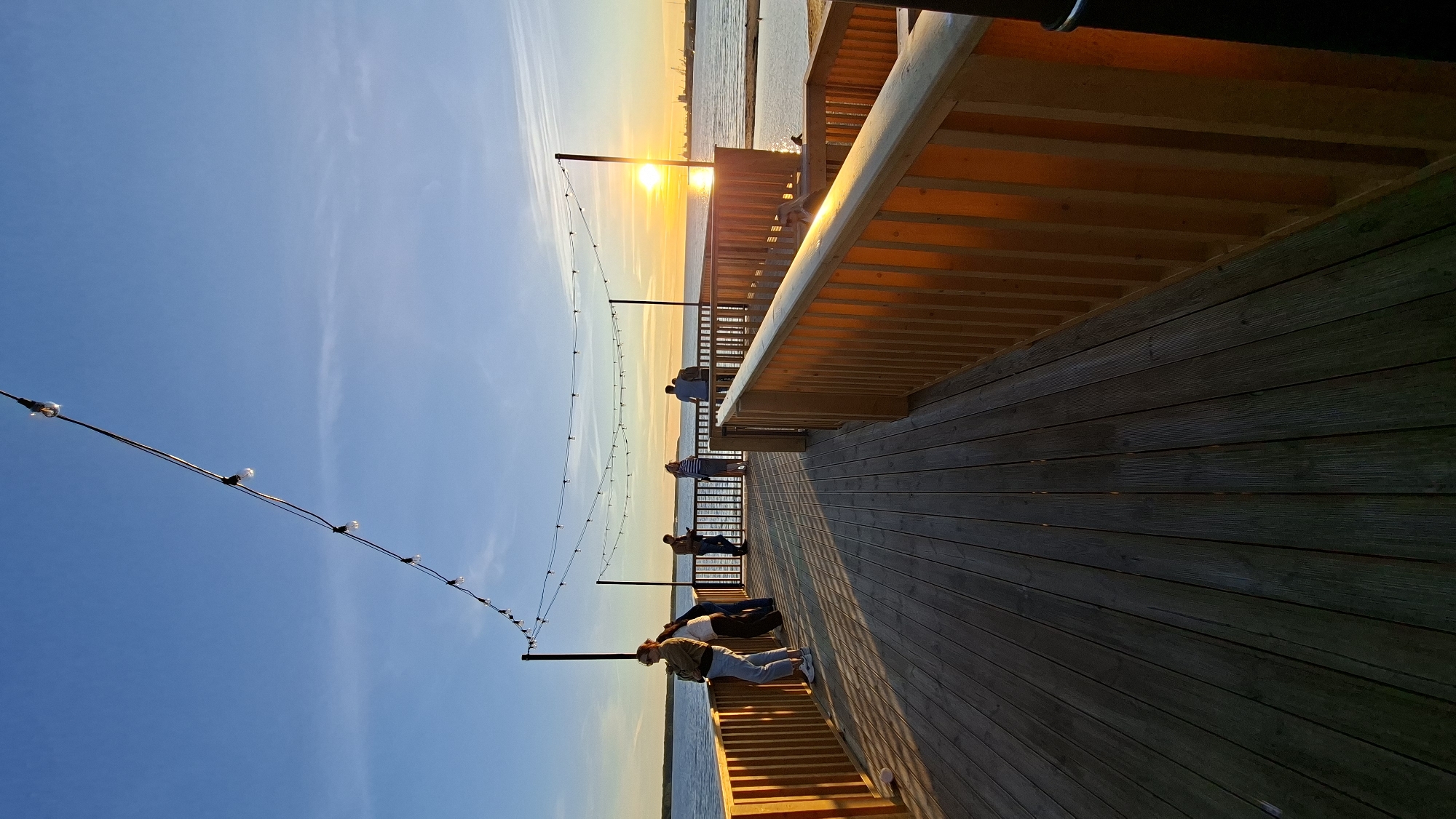 This image depicts a wooden boardwalk overlooking a body of water at sunset. The sky is clear blue with hints of orange and yellow as the sun sets on the horizon, reflecting on the water. String lights are hung above the boardwalk, adding a festive ambiance. Several individuals are visible on the boardwalk, some standing and looking out at the water, while others appear to be walking or conversing. The boardwalk has wooden railings, and the wooden planks of the deck are clearly visible. In the distance, industrial structures and a large body of water suggest a coastal or port area. Based on the presence of industrial port structures and the overall aesthetic, this location is likely the Tallinn Maritime Museum or a similar area along the waterfront in Tallinn, Estonia.