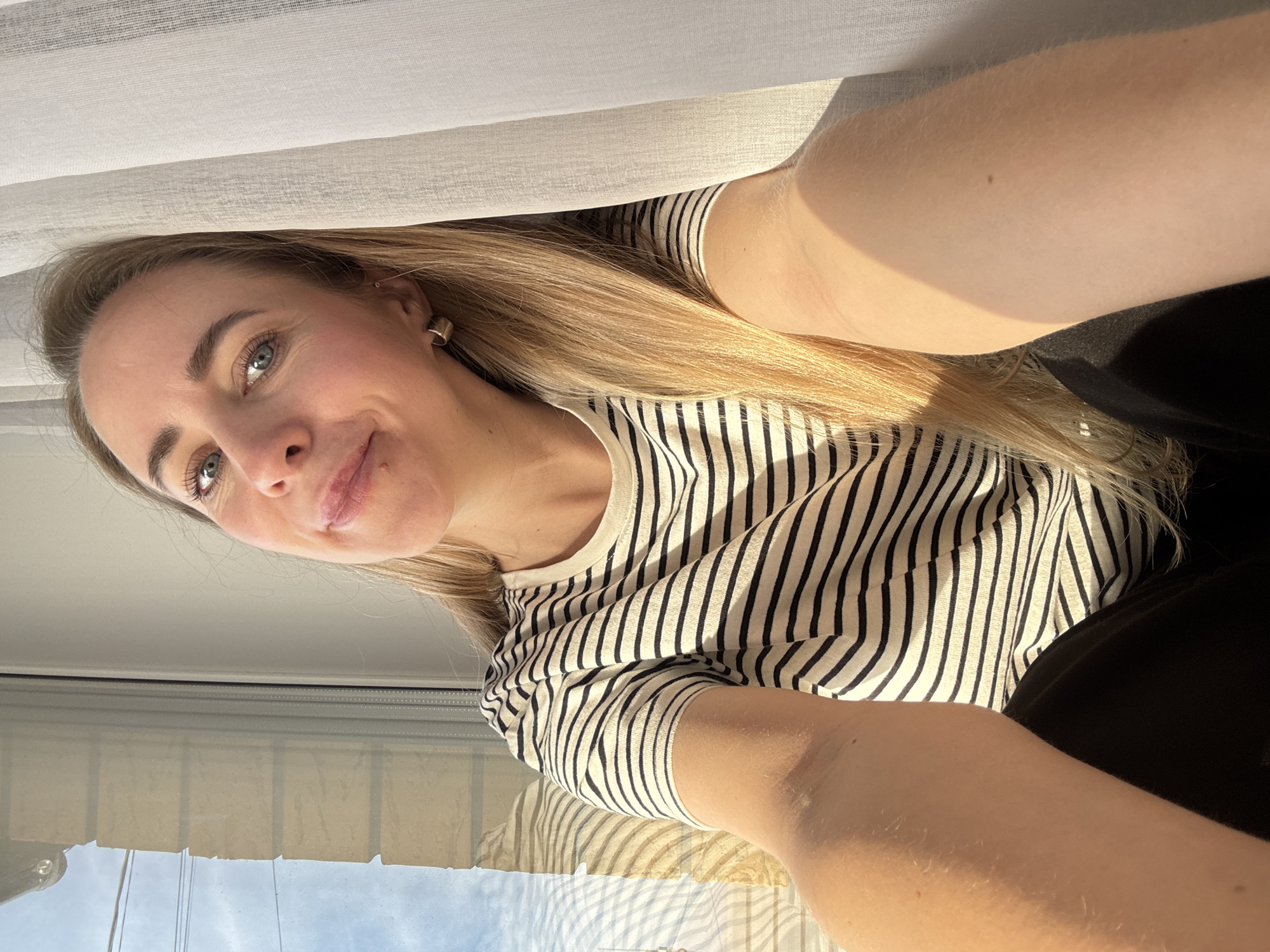A young woman with long blonde hair and light blue-green eyes is captured in a vertically oriented selfie. Her head is tilted, and she displays a soft, pleasant smile, looking slightly upwards and to the side. She wears a cream or off-white t-shirt with thin black horizontal stripes and a simple gold hoop earring in her visible ear.
The scene takes place indoors, likely in Chaikovsky, Russia, near a window. Bright natural sunlight streams in, illuminating her face and hair and casting gentle shadows. A light-colored sheer curtain or blind occupies the upper portion of the frame. Through the lower section of the window, a clear blue sky is visible, along with reflections of buildings or structures, suggesting an urban or suburban setting. Her arms are extended towards the camera, with a glimpse of a dark, possibly black, garment visible on the lower right side of the image. The overall mood conveyed is one of calm and contentment on a sunny day. No text is visible.