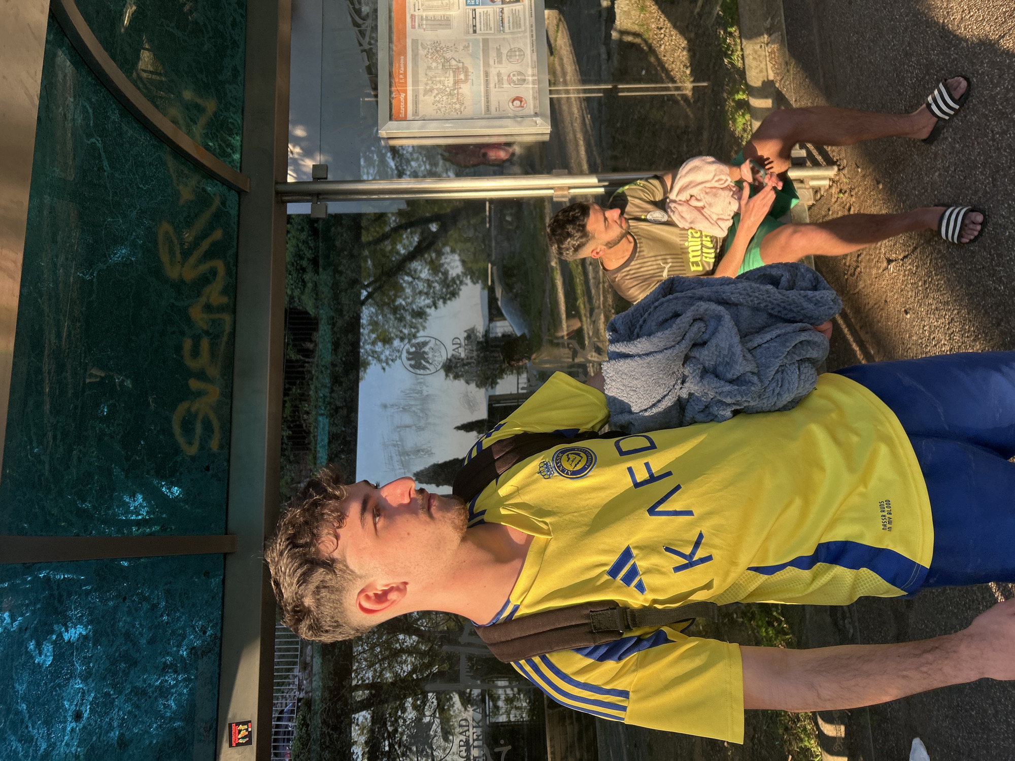 Two men are at a bus stop in Rijeka, Croatia. One man, in a yellow and blue soccer jersey, stands facing left, with a dark backpack, while the other man is sitting on the bus stop bench, wearing a brown and green shirt and green shorts, with a pink towel on his head. He is looking at his cell phone. He is wearing a pair of black and white Adidas slides. A blue sweater is on the bench next to him. The bus stop has a clear glass back with information on it. The green marble glass on the side of the bus stop has the word 'SUENTLY' in yellow writing. A sign underneath the glass contains the words, 'GRAD LIEKA.' The scene appears to be outside, with trees and a building reflecting in the bus stop glass.