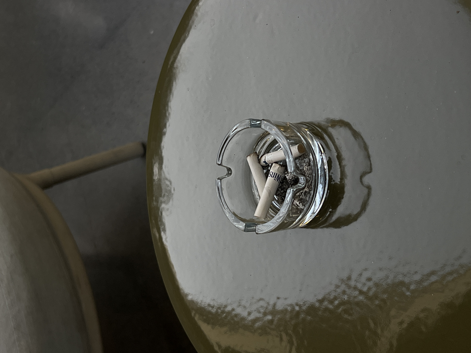 An ashtray filled with cigarette butts and ash rests on a glossy, olive-green table. The ashtray is made of clear glass and has three indentations for cigarettes. The table is round and reflects light, showing some blurred shapes in the background that suggest a seating area with furniture. The scene appears to be outdoors, possibly in Tel Aviv, Israel, given the context provided, and suggests a moment of relaxation or a break. There are no people visible in the image.